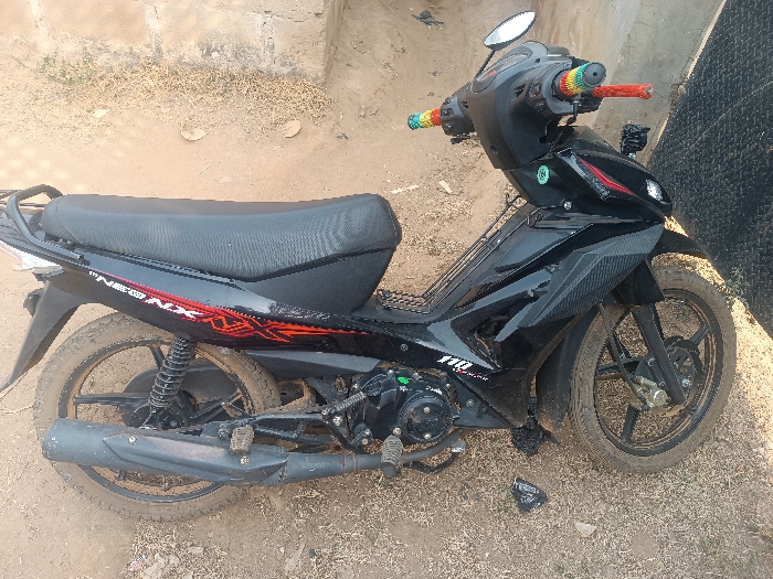 The scene, located in Oye, Nigeria, displays a black underbone motorcycle parked on a dry, light brown dirt ground with sparse, dried vegetation. The motorcycle is viewed from its left side. It features black body panels with red and white graphic decals on the side fairing, including text such as "NEW NX" and "110". Both the front and rear tires and black alloy wheels are heavily coated with light brown dirt. The handlebars are fitted with multicolored (rainbow-patterned) grips on both sides, and a single silver rearview mirror is present on the left handlebar. A black exhaust pipe extends along the lower left side, also covered in dirt. A small, crumpled black plastic bag is visible on the ground near the motorcycle's front wheel. In the background, a light-colored, possibly concrete or brick wall is partially visible on the left, and a dark, textured surface, potentially a metal gate or wall, is visible on the right. No individuals are present, and no actions are depicted.