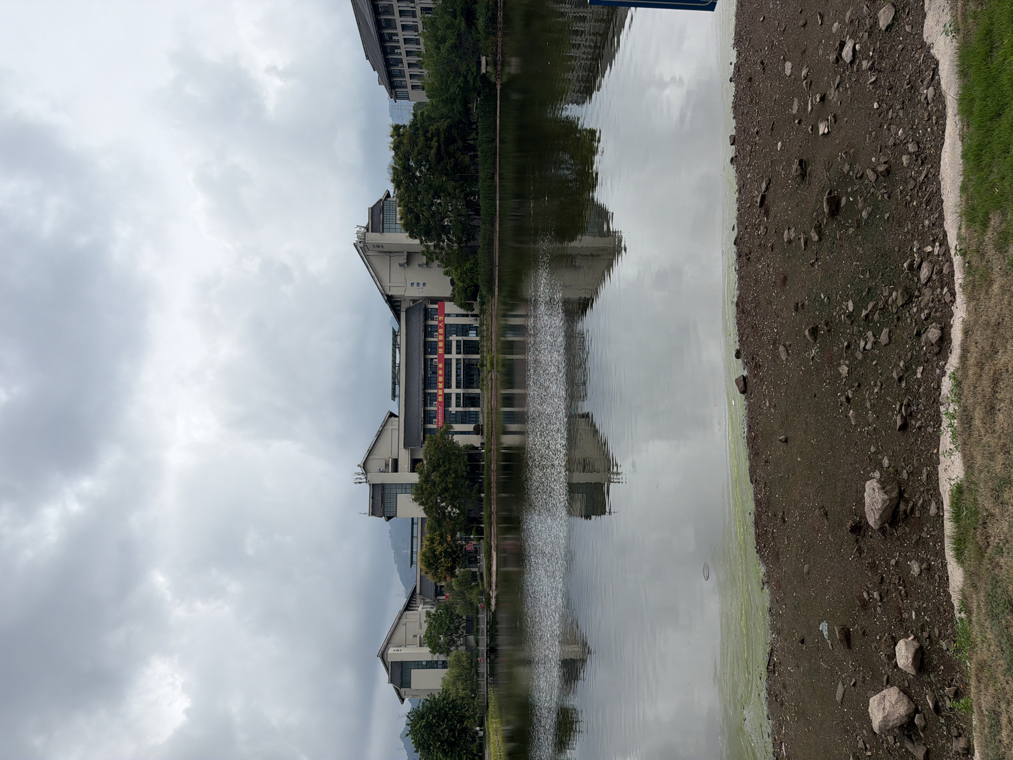 A wide-angle, eye-level shot captures a tranquil scene on a cloudy day at a body of water, likely a lake or reservoir, in Jiangjin, China. In the foreground, a muddy bank is strewn with various-sized stones, some grass, and patches of green algae bloom floating on the water's edge. The water itself is calm and reflects the overcast sky and the buildings on the opposite shore. A band of lighter green algae is visible across the water's surface in the middle distance.
Dominating the background is a complex of modern, multi-story buildings, exhibiting a mix of architectural styles. These structures appear to be institutional or commercial, with some featuring pitched roofs and others flatter ones with visible antennas. One building in the center has a prominent red banner with Chinese characters hanging across its facade. The buildings are surrounded by mature trees, adding a touch of greenery to the scene. The reflection of the buildings and trees is clearly visible in the still water. Beyond the buildings, rolling hills or mountains can be seen in the distance under a heavily clouded sky. The overall atmosphere is subdued and slightly melancholic due to the cloudy weather. No people are visible in the image.