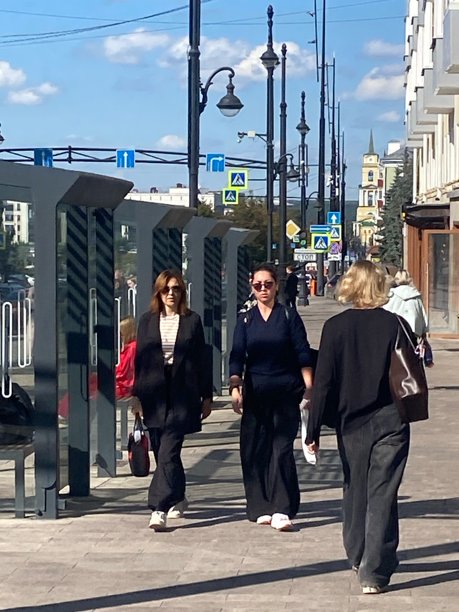 The image depicts a street scene in Perm, Russia, likely on a sunny day. Three women are walking along a paved sidewalk. The woman in the foreground on the right wears a black long-sleeved top and dark pants, carrying a dark brown shoulder bag. She has blonde hair and is walking away from the camera. To her left, another woman in a dark blue v-neck top and wide-leg dark pants walks towards the camera, wearing sunglasses. A third woman, on the far left, is seen from the chest down, wearing a dark blazer, striped shirt, and dark pants. She is also walking away from the camera, carrying a dark bag with red accents.
The sidewalk is bordered by modern, glass-paneled bus shelters with dark frames on the left. Beyond the shelters, a street with parked cars is visible. Across the street, a building with white architectural elements and balconies lines the right side of the frame. Further in the background, a church with a distinctive spire is visible, along with other buildings and trees. Traffic signs, including pedestrian crossing signs and directional arrows, are mounted on poles along the street. The sky is blue with scattered white clouds. The overall impression is a typical urban environment on a clear day.