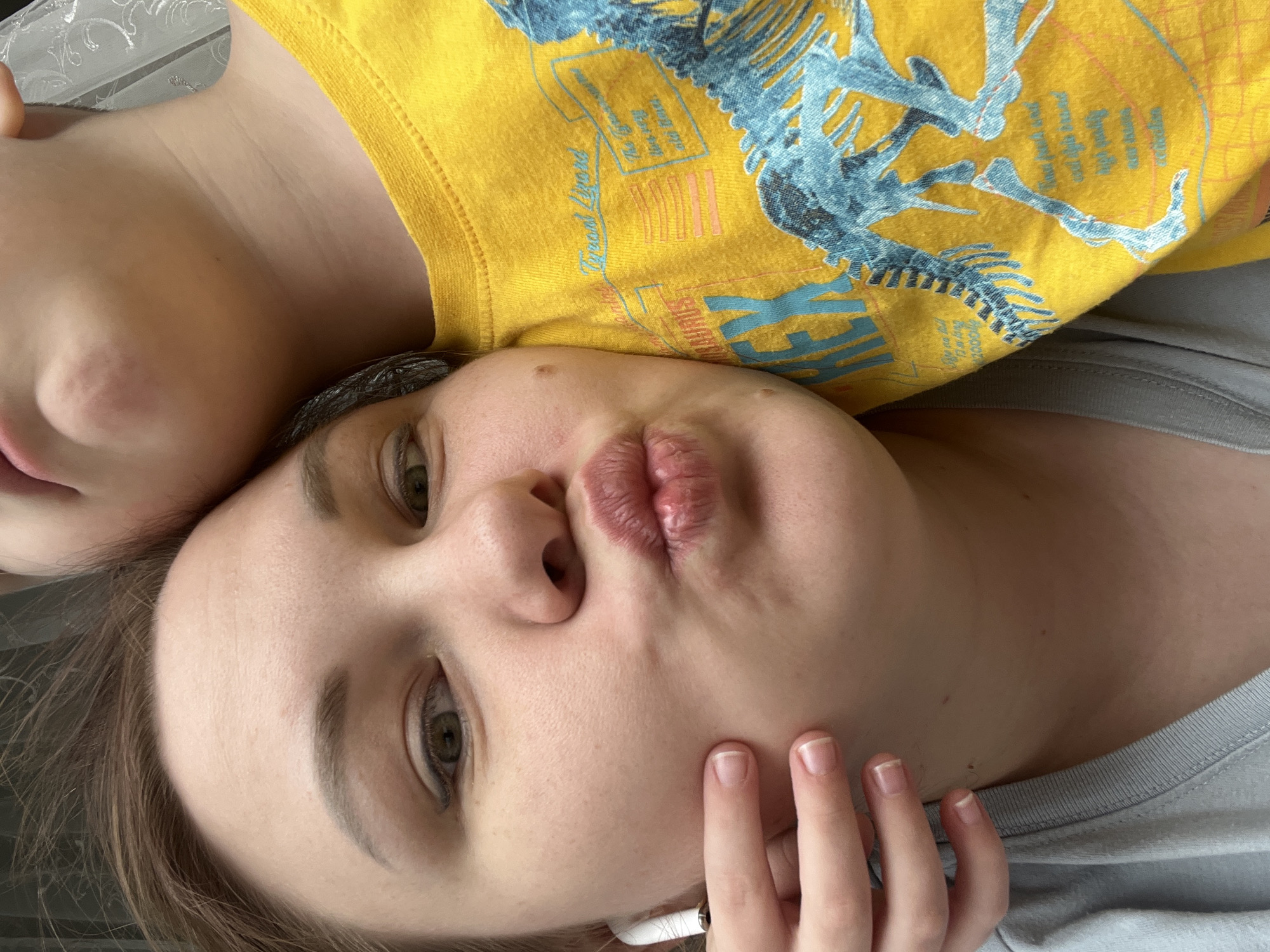A close-up, face-level photograph shows a woman with light skin, light brown hair, and green eyes lying next to a child. The woman is oriented at the bottom, with the child above. The woman has pursed lips and her fingers are near her face. She is wearing a gray shirt and an AirPod is visible in her ear. The child is wearing a yellow shirt with a blue dinosaur graphic. The background is blurred. The location in Usman, Russia is not identifiable from the image.