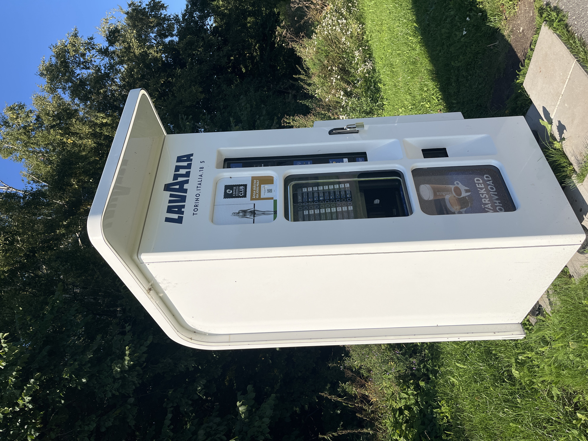 A white outdoor vending machine, branded "LAVAZZA TORINO.ITALIA.18 5", is situated outdoors on a sunny day. The machine is in a grassy area with trees and foliage in the background. It displays various options for beverages, including a picture of coffee and the text "VÄRSKED KOHVIOAD", indicating fresh coffee beans. A padlock is attached to the right side of the machine. The machine is positioned on concrete slabs. The environment suggests a public outdoor space, possibly a park or rest stop within the city of Kohtla-Järve, Estonia.