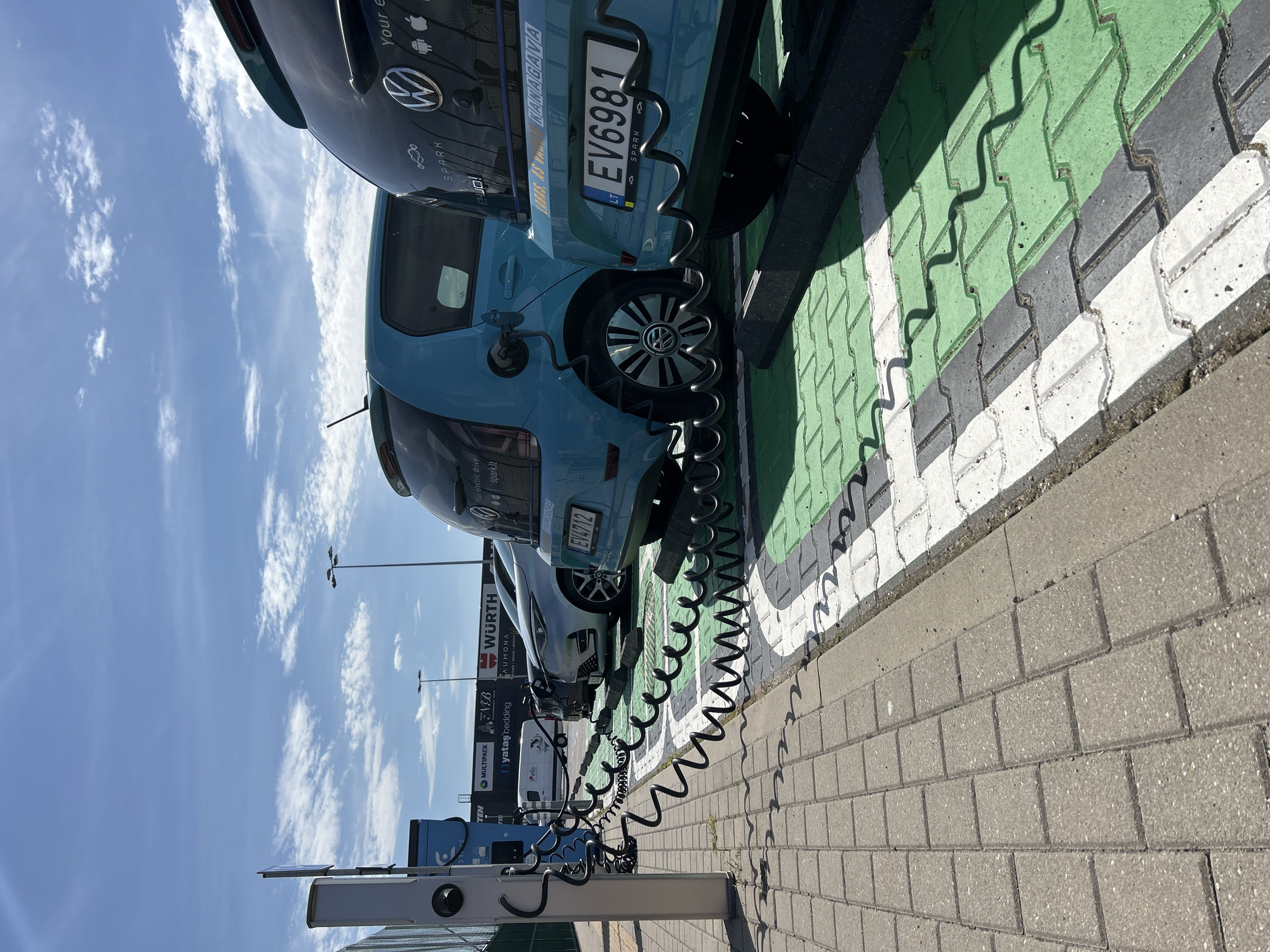 In Vilnius, Lithuania, an outdoor scene captures an electric vehicle charging station bathed in daylight. Several electric cars, including a blue Volkswagen ID.3 and a darker vehicle, are parked in designated charging spots marked with green and white pavement. Charging cables, coiled and extending from charging units on a tall white pole, are connected to the vehicles. The background reveals a strip of buildings with visible signage, including "WURTH" and "MULTIPACK," suggesting a commercial or industrial area. The sky above is a clear blue, punctuated by scattered white clouds. The foreground shows a pedestrian walkway paved with interlocking gray concrete blocks.