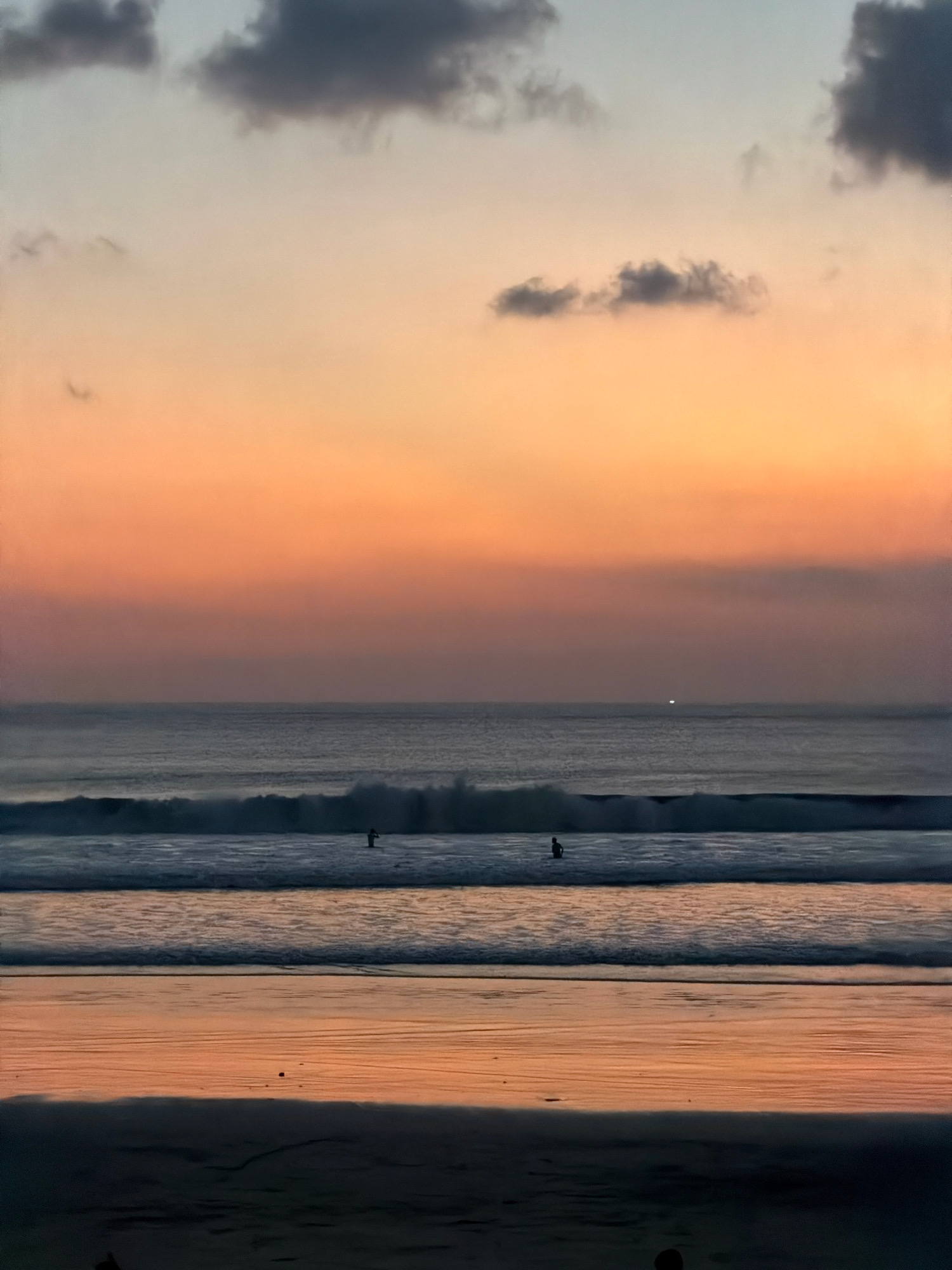 A serene sunset scene at Kuta Beach, Indonesia, captures the tranquil beauty of the ocean at dusk. The sky transitions through a gradient of soft oranges, pinks, and blues, with scattered dark clouds adding depth to the horizon. Two surfers are visible in the distance, standing in the shallow water, their silhouettes outlined against the gentle waves. The ocean itself is a calm expanse of deep blue, with small waves lapping at the shore. The wet sand in the foreground reflects the warm hues of the sunset, creating a mirror-like effect. The overall atmosphere is peaceful and contemplative, inviting the viewer to soak in the natural splendor of the moment.