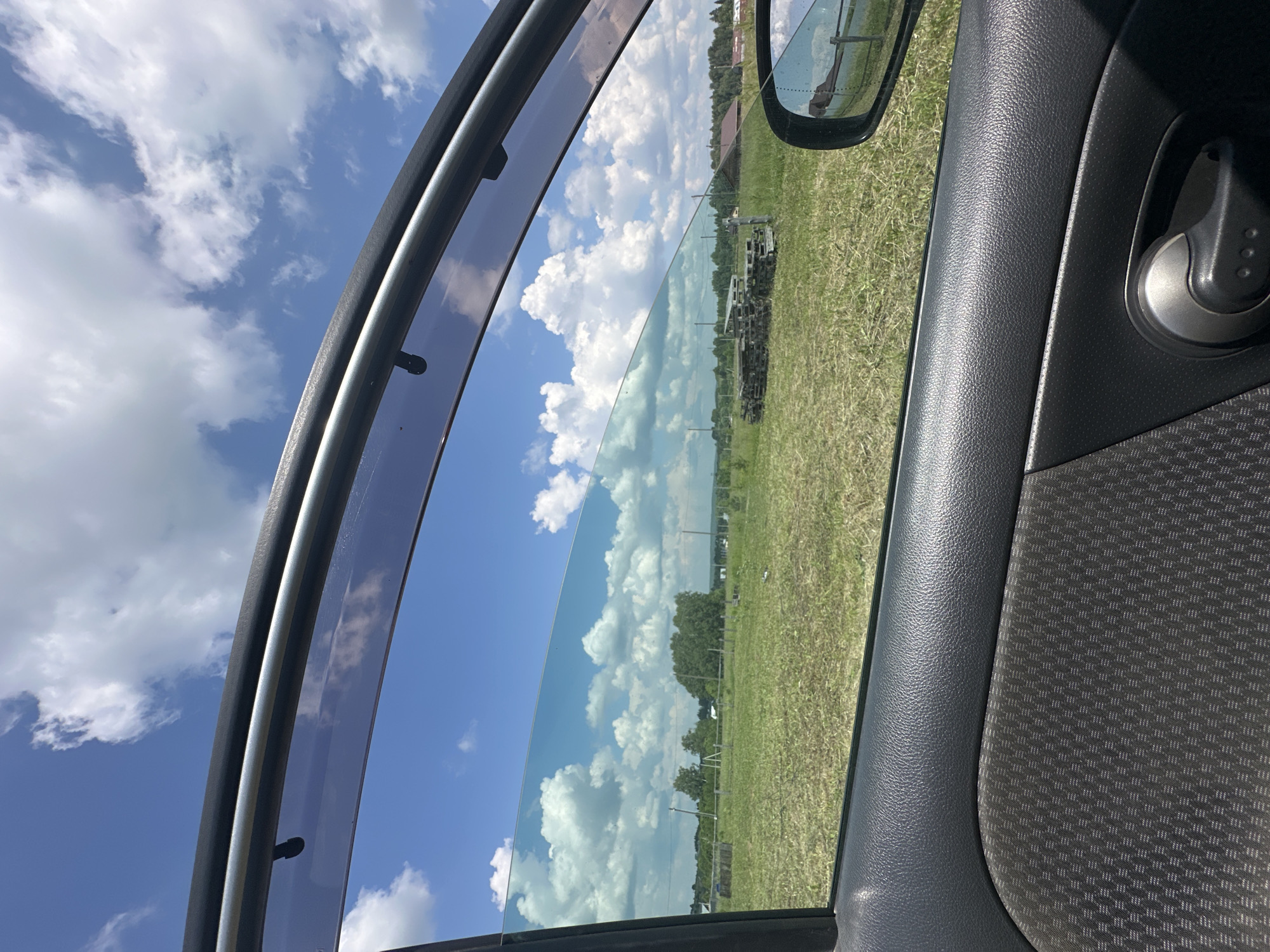 The image is a view from inside a car, looking out the open window at a rural landscape. The foreground shows the interior of the car door, with dark grey textured plastic and fabric panels, and a chrome door handle. Outside the car, a sunlit field of green grass and some dry stalks stretches into the distance. In the middle ground, there are piles of wooden pallets and some scattered farming equipment. Further back, trees and small houses suggest a village. The sky is a bright blue with fluffy white clouds. A side mirror is visible on the right, reflecting a fence and a portion of the sky. The location is identified as Malyi Bukor, Russia.