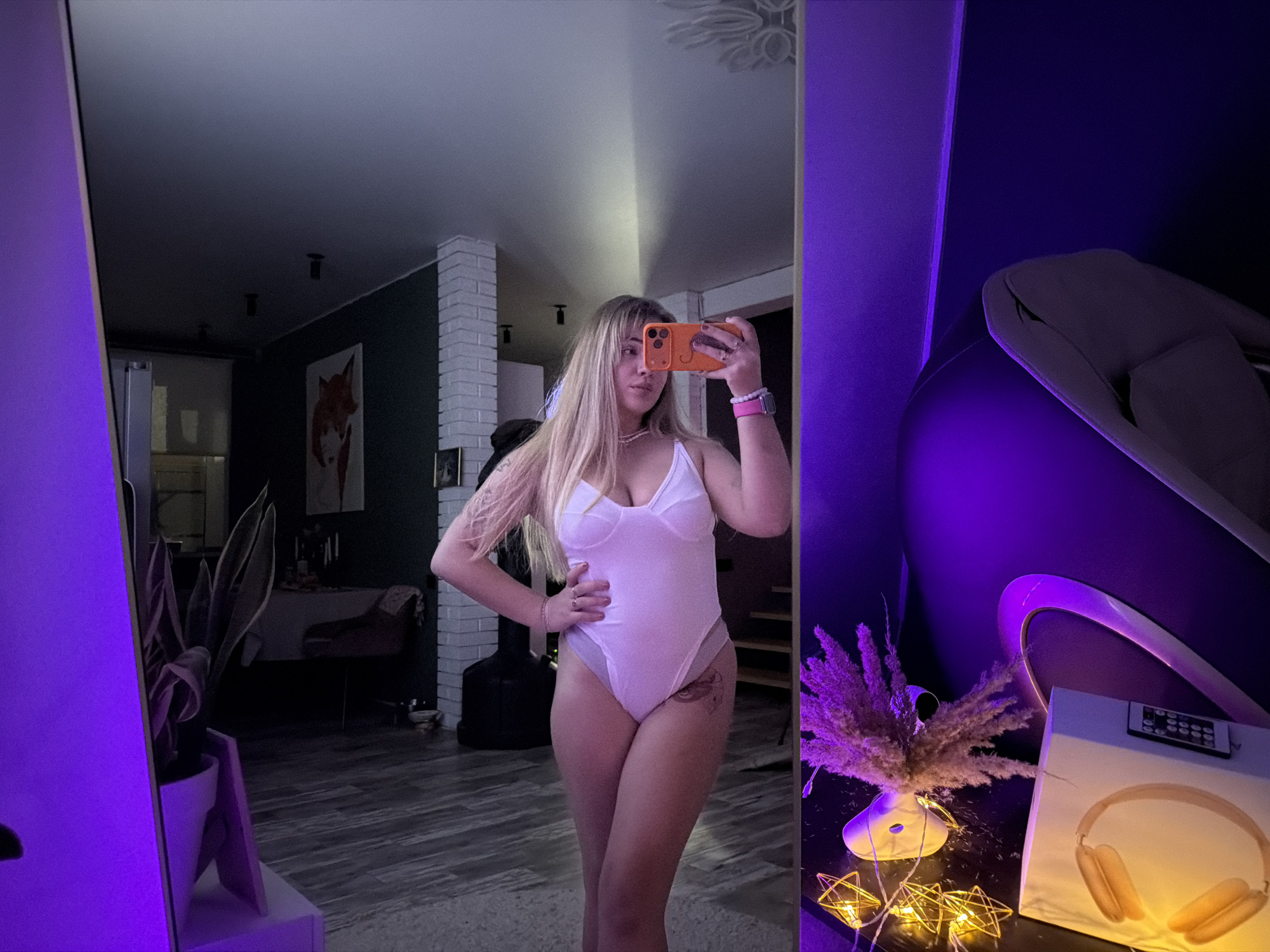 A young woman stands in front of a full-length mirror, holding a cell phone up to take a picture of herself. She is wearing a white bodysuit and a necklace. Her hair is blonde and shoulder-length. The background includes a living room setting with a dining table, a piece of art on the wall, and decorative plants. There is also a piece of modern furniture with purple lighting on the right side of the image. The location is in Большая Шильна, Russia.