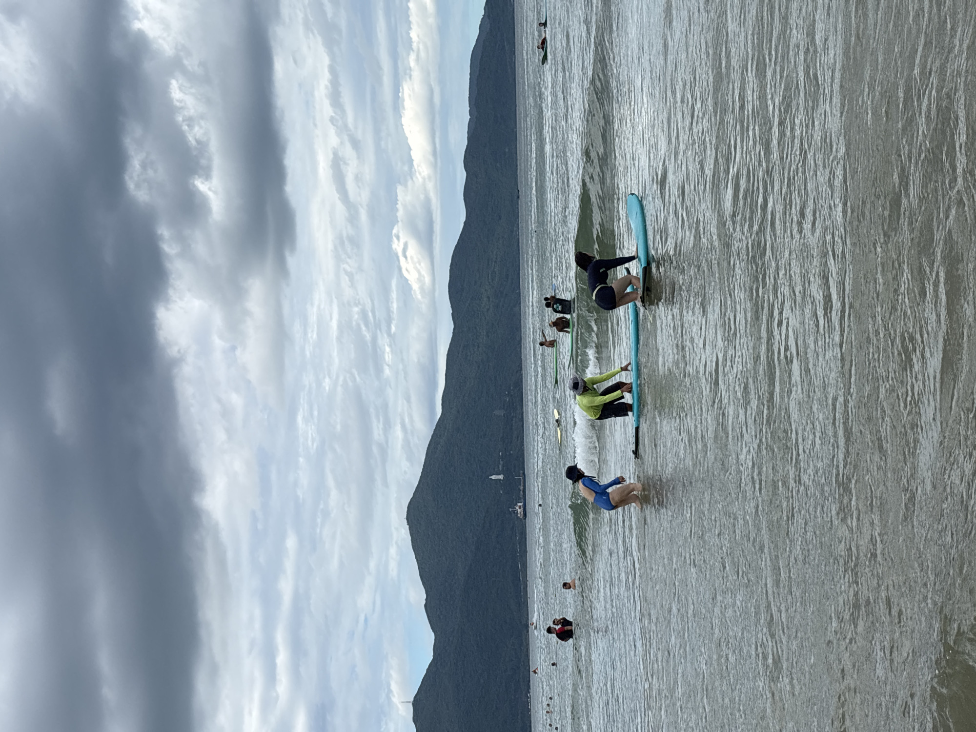 This image captures a scene at Mỹ Khê Beach in Đà Nẵng, Vietnam, on a cloudy day. The water is calm with gentle waves, and the sky is filled with dark, moody clouds. In the foreground, several people are engaged in water activities. Two individuals are on paddleboards, with one wearing a blue long-sleeved rash guard and the other in a green long-sleeved top and dark shorts. A person in a black outfit is kneeling on a blue paddleboard. Further out in the water, other people are visible swimming and standing in the shallow waves, some with surfboards. In the background, lush green mountains form a dramatic backdrop to the coastline, with a visible tower atop one of the peaks.
