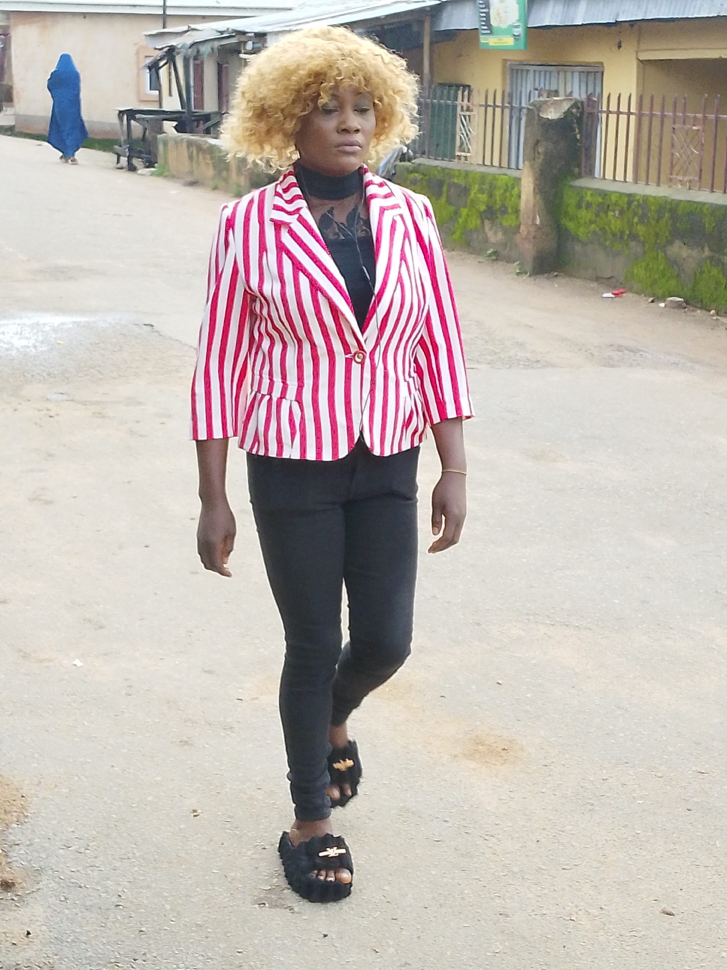 A woman with blonde, curly hair walks down a paved road in Jos, Nigeria. She is wearing a red and white striped blazer over a black top and black skinny jeans. She has on black fluffy slides with gold accents. Her left hand is by her side, and her right hand is slightly forward. In the background, a person wearing a blue headscarf and robe walks away from the camera on the left side of the road. Buildings and a metal fence are visible along the right side of the road, with moss growing on a stone wall in front of them. The scene appears to be during the daytime.
