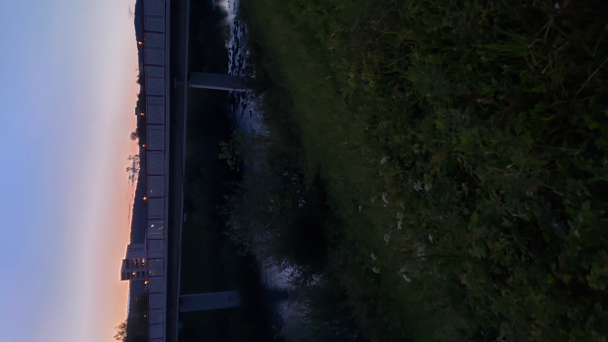 The image captures a twilight scene in Prešov, Slovakia, featuring a concrete bridge spanning a river. The bridge has a railing with red accents and lights. On the far bank of the river, a modern building with lit windows is visible behind a line of trees. The river itself appears calm with some visible rocks. The foreground is dominated by dark, grassy vegetation, with a few scattered white flowers. The sky above displays a gradient of colors, transitioning from a deep blue at the top to softer pinks and oranges near the horizon. The overall atmosphere is serene and slightly dim, indicative of the early evening.