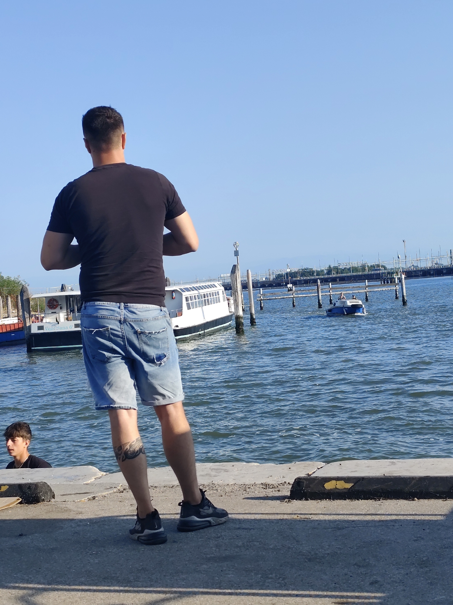This is a photograph of a man standing on a promenade in Venice, Italy, facing the water. He is wearing a black t-shirt and ripped denim shorts. Behind him, on the water, are several boats, including a large white passenger ferry labeled "Veneziana 6". In the distance, a smaller blue and white motorboat is visible with two people on board. The sky is clear and blue, and the water is choppy. Another young man is visible on the left side of the image, looking out at the water. The promenade is made of concrete and has a black bumper along the edge.