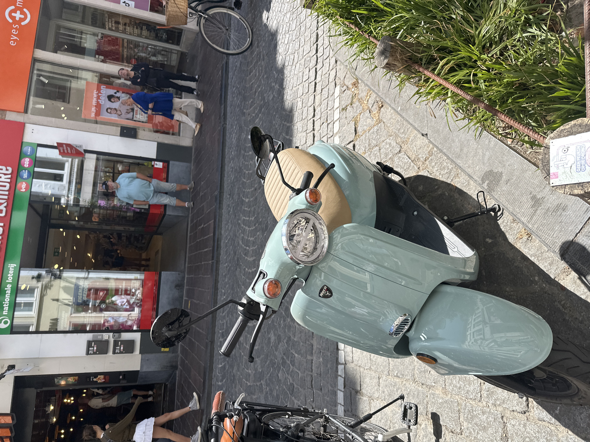 In Bruges, Belgium, on a sunny day, a light blue retro-style scooter is parked on a cobblestone street. The scooter has a tan seat and a silver emblem on its front. To the left of the scooter, a bicycle is parked, its rear wheel visible. In the background, a store with a green awning that reads "nationale loterij" and "more" is open. Several people are seen near the store entrance and on the sidewalk. One person in a light blue shirt and grey shorts stands near the store. Another person in a blue jacket and white pants walks away from the camera. The street is lined with buildings, and a section of green grass is visible in the foreground, with wooden posts and a rusty metal rod.