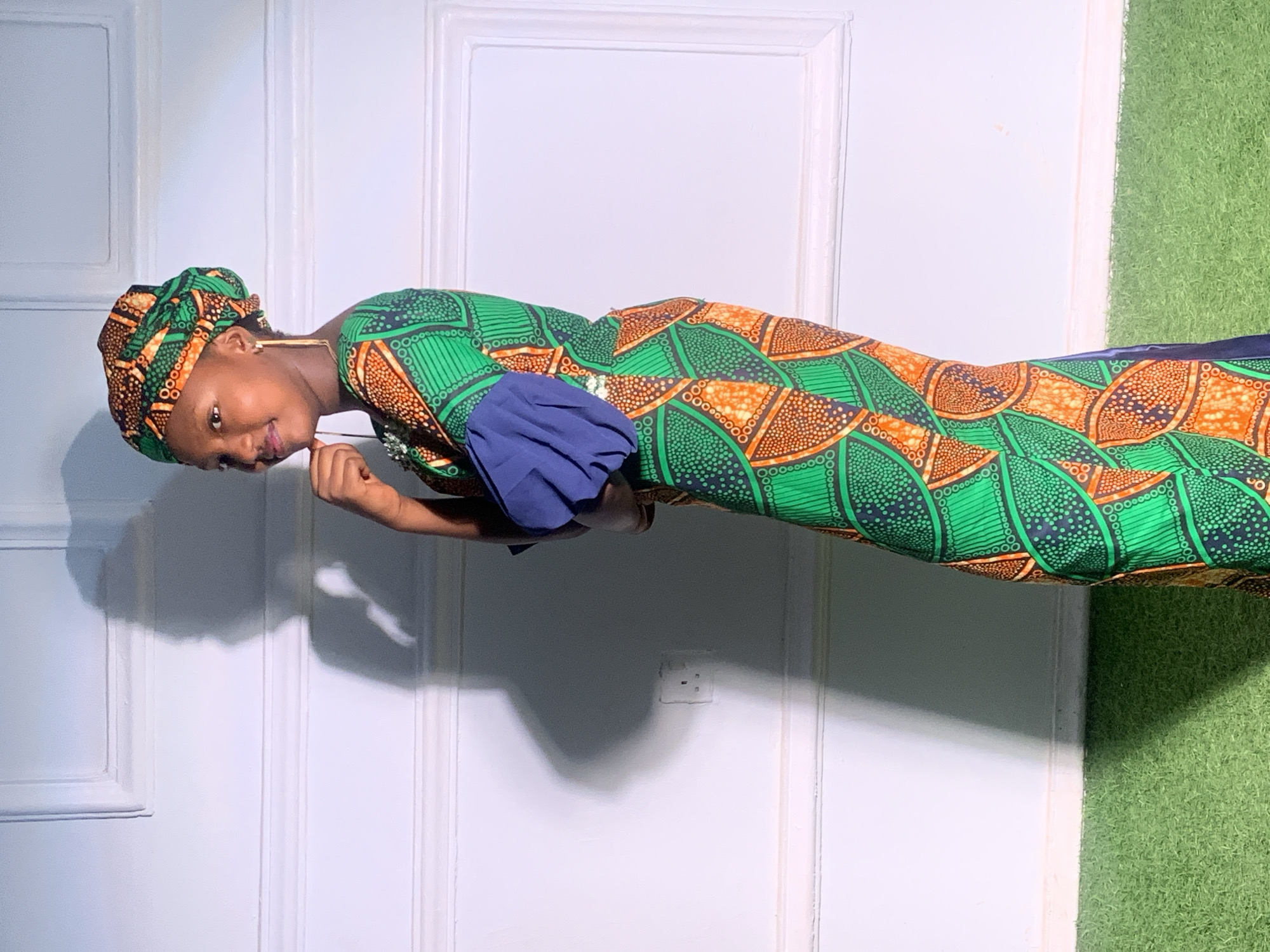 A woman is posing in front of a white paneled door and a patch of artificial grass. She is wearing a brightly patterned, green and orange dress with a matching head covering. The dress has a blue sleeve. The woman is smiling slightly and has her hand near her chin. A power outlet is visible on the wall behind her. The location is likely indoors, but there are no indicators that it is in Barakin Akawo, Nigeria.