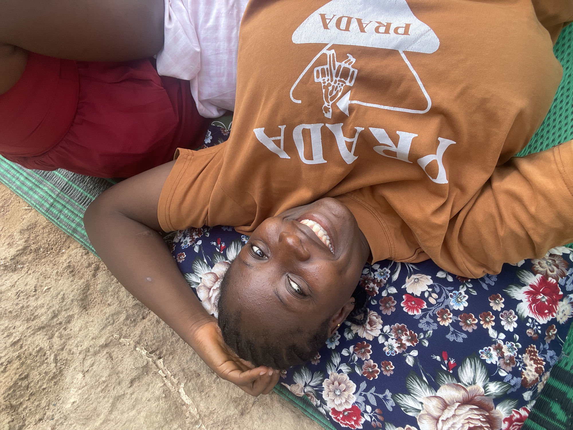 A young woman lies on her back on a floral-print pillow placed on a woven green and white mat, which is itself on sandy ground. She is smiling broadly, looking directly at the camera. She wears an orange t-shirt with the word "PRADA" printed in white. She also wears a red garment and a white garment. The setting appears to be outdoors in Akwanga, Nigeria, evidenced by the location metadata.