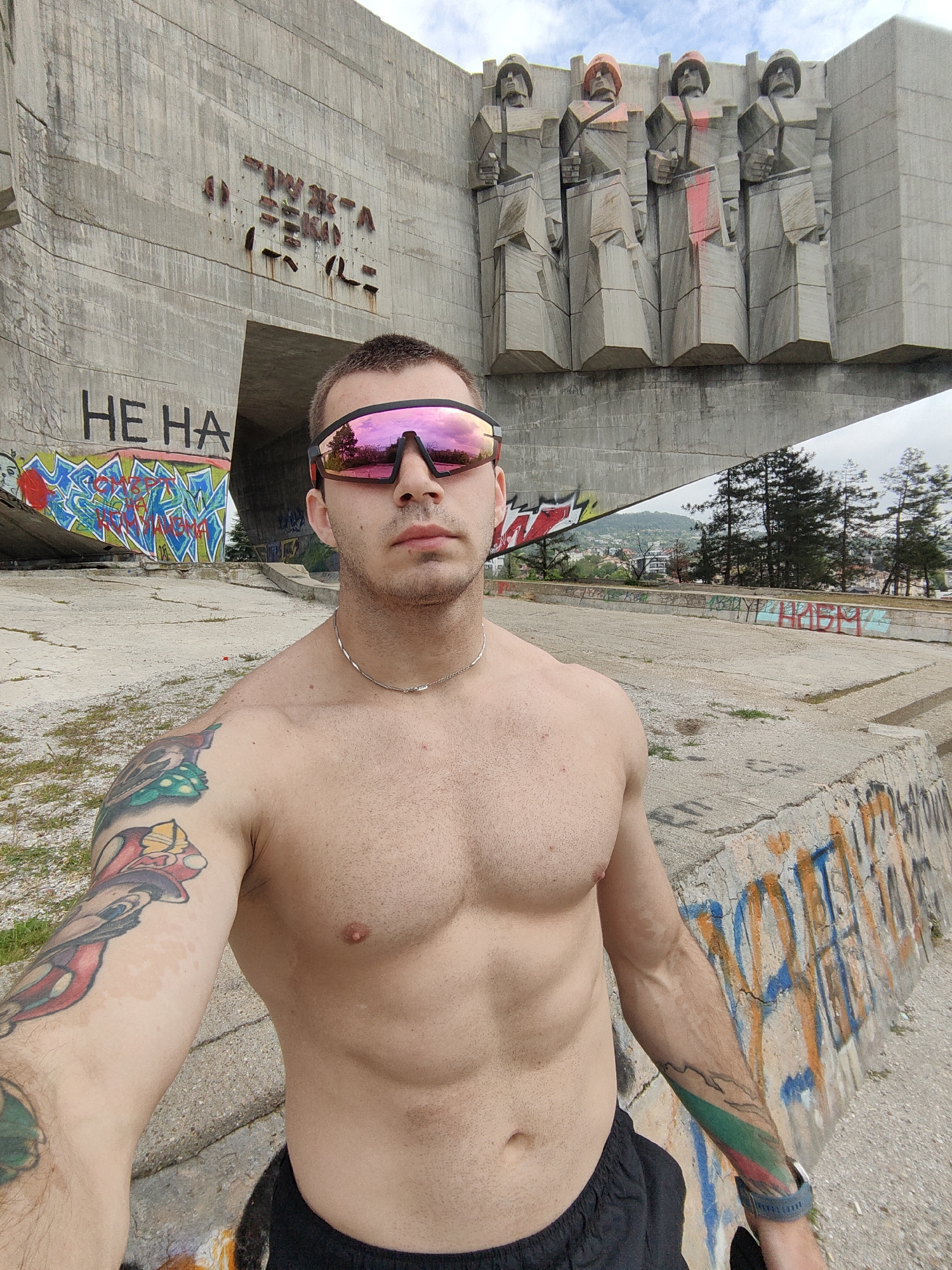 A shirtless man wearing sunglasses and a necklace is taking a selfie in front of the Monument of the Bulgarian-Soviet Friendship in Varna, Bulgaria. The monument is a large concrete structure with graffiti and four Soviet figures near the top. The surrounding area is also covered in graffiti. The man has tattoos on both arms. The sky is cloudy.