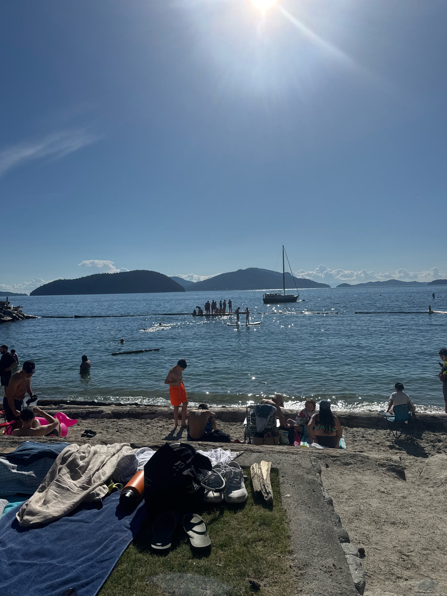 The image depicts a sunny day at a beach in Lions Bay, Canada, with the clear blue sky and bright sun dominating the upper portion of the frame. The water is calm, with a few small waves lapping at the shore. A sailboat is anchored in the distance, and several people are visible in the water and on the beach.
On the beach, several individuals are enjoying the weather. In the foreground, a collection of beach items is laid out on towels and grass, including a black backpack, a water bottle, and various articles of clothing. A few pairs of shoes and flip-flops are also scattered around.
Further back on the sandy shore, people are seated on chairs or lounging on towels, some interacting with each other. The water is a central feature, with several people swimming or wading. A group of individuals is standing on a floating platform or dock-like structure in the water, while others are seen on paddleboards in the distance. The shoreline is a mix of sand and some rocky areas.
The background features a picturesque view of islands and mountains under the bright sun, typical of a coastal Canadian landscape. The scene conveys a relaxed, recreational atmosphere on a warm summer day.
