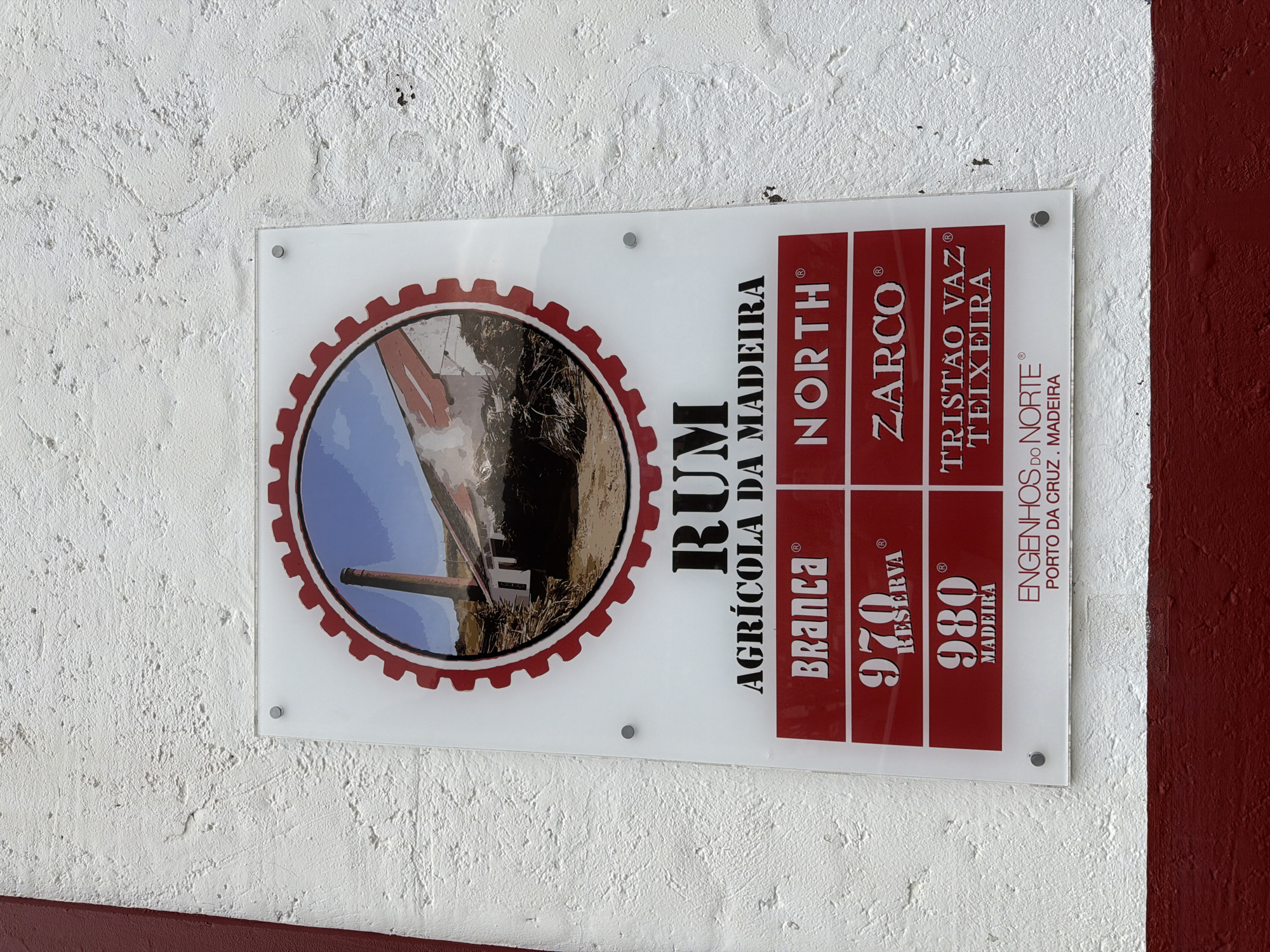 The image shows a sign for "Rum Agricola da Madeira" located in Porto da Cruz, Machico, Portugal. The sign is rectangular, mounted on a wall, and made of a clear material with white and red lettering. It features an image of a sugar cane mill. The sign lists different types of rum: "Branca," "970 Reserva," and "980 Madeira," and geographical names associated with rum production: "North," "Zarco," and "Tristão Vaz Teixeira." The sign also includes the name of the rum producer: "Engenhos do Norte." The wall behind the sign is painted white with a textured surface, while a red section is visible at the right edge.