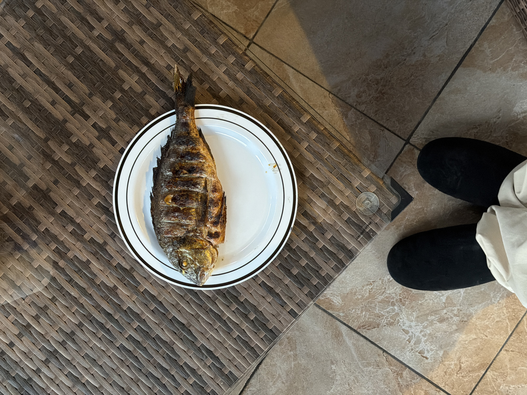A cooked fish is centered on a white plate with a black border, placed on a brown wicker table. Adjacent to the table, on a tiled floor, are two black slippers and part of a white garment. The image appears to have been taken at an indoor location in Новоглаголево, Russia.