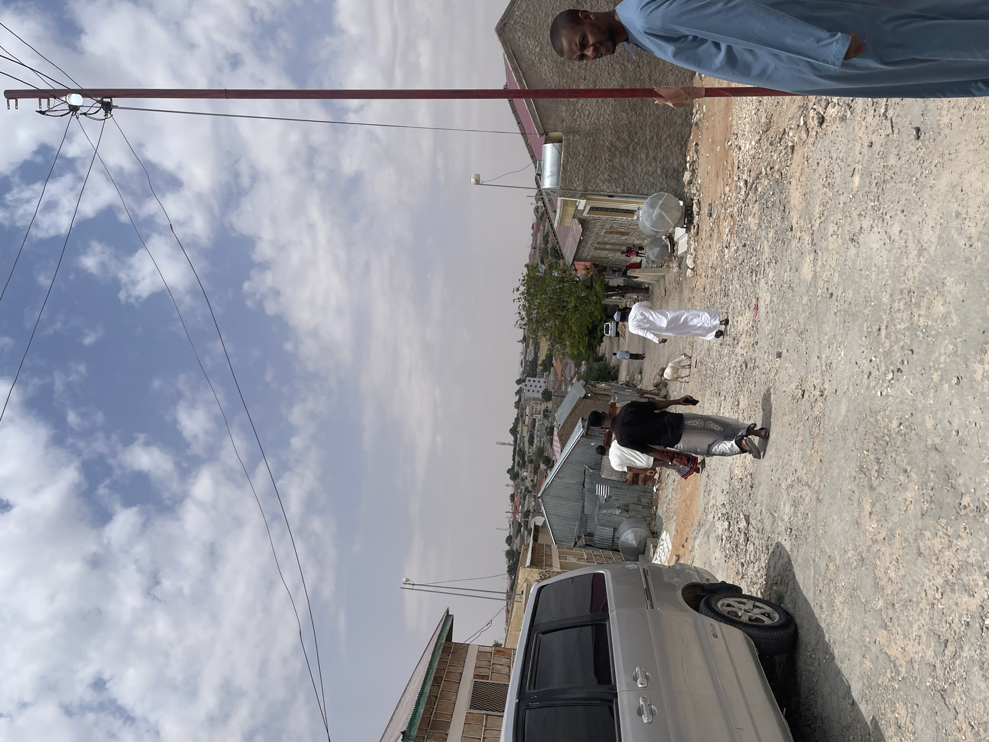 An eye-level, medium shot captures a dusty street scene in Hargeisa, Somaliland, under a partly cloudy sky. On the right, a man in a blue robe stands partially facing the camera, his expression unreadable. A red utility pole with wires and a lit bulb stands in the foreground. The street is unpaved and rocky, with a silver van parked on the left. Several people are walking down the street, including a man in a black shirt and patterned pants, another in a white robe, and a few others further in the distance. Goats wander near the people. Buildings with corrugated metal roofs and concrete walls line the street, suggesting a mix of residential and commercial structures. The background shows a dense collection of buildings stretching into the distance, characteristic of a city. The overall impression is one of everyday life unfolding in an arid urban environment.