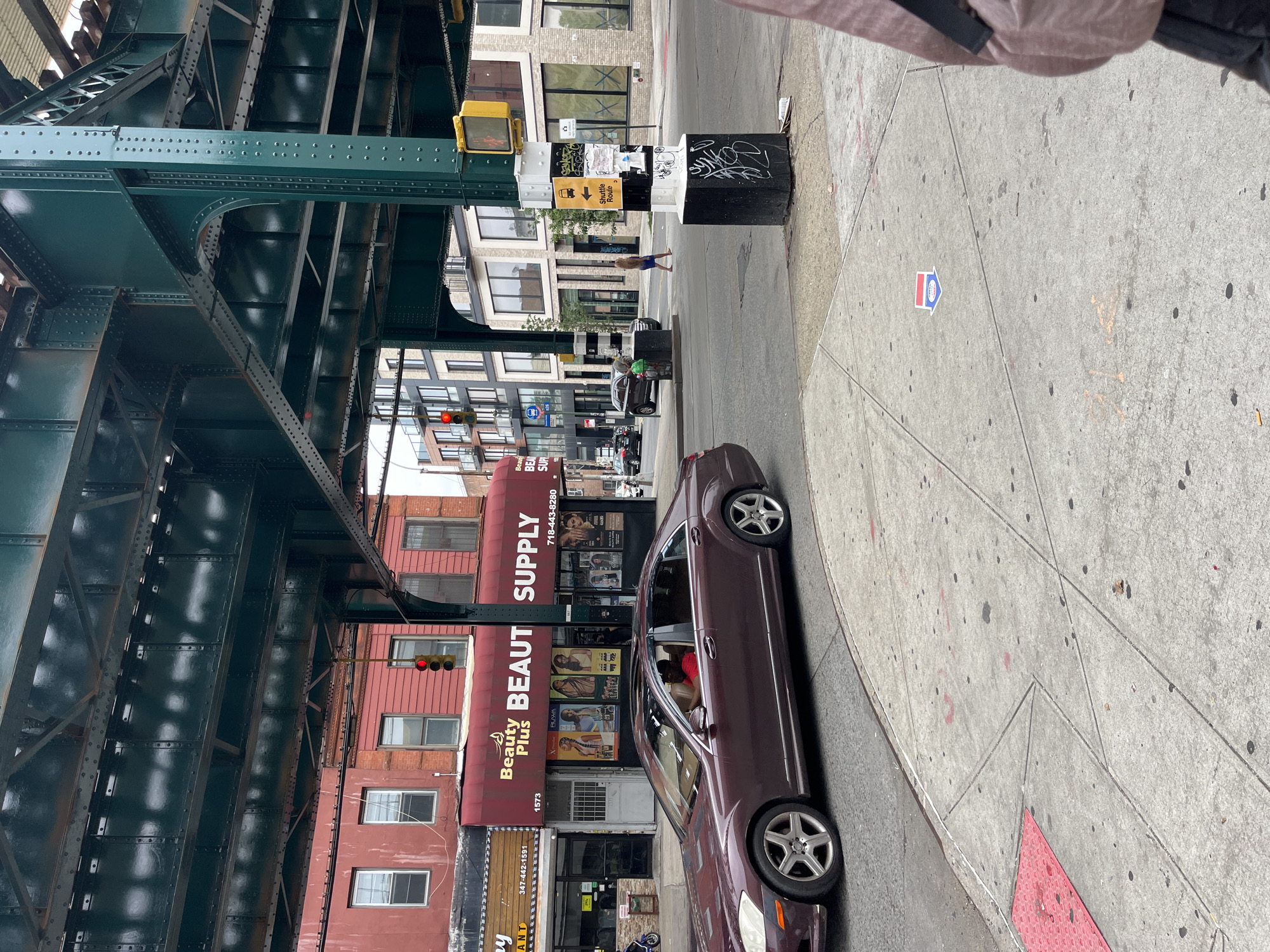 The image depicts an urban street scene in New York City, United States of America, under an elevated train line. A maroon Mercedes-Benz sedan is driving on the road in the foreground. On the sidewalk to the left, a business called "Beauty Plus Beauty Supply" is visible with posters displayed on its facade. A traffic light is red, indicating that vehicles should stop. Further down the street, modern buildings with large windows are present, alongside older red brick structures. Pedestrians are visible on the sidewalk, and a woman in a blue dress is crossing the street. The environment includes concrete sidewalks, asphalt roads, and the substantial green metal infrastructure of the elevated train line overhead. Signs for "Shuttle Route" and other pictograms are attached to a utility pole. There is graffiti on the base of the utility pole, and various pieces of trash are scattered on the ground.