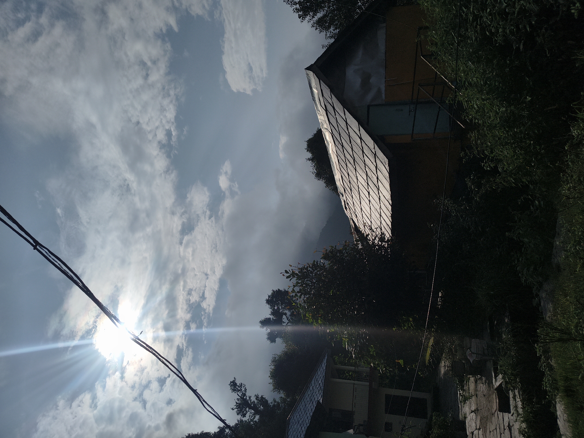 The image depicts a rural scene in Haini, India, featuring a small, rustic house with a tiled roof. The sun is bright and casting rays through clouds in the sky, creating a lens flare effect. Power lines are visible in the foreground. The surrounding environment is lush with trees and vegetation, suggesting a natural or mountainous setting. The house appears to be made of yellow and blue painted materials with a metal roof. There are no people visible in the image. The overall atmosphere is one of natural beauty and tranquility, with the sun's rays adding a dramatic element to the scene.