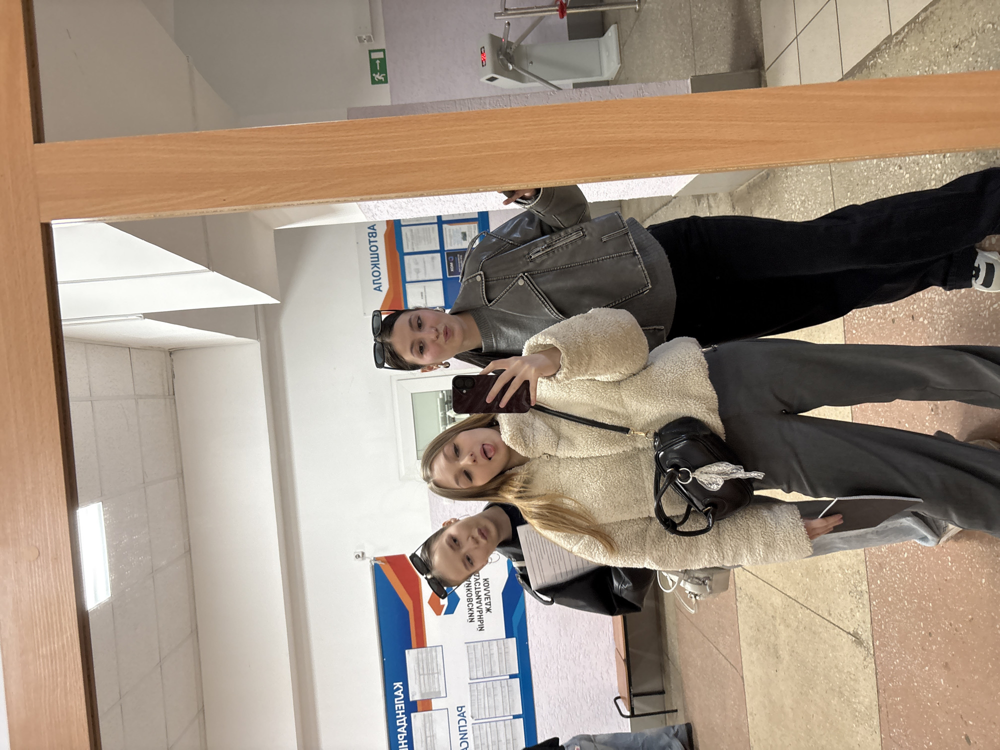 Two young women are captured in a mirror selfie, taken indoors, likely in a public building or educational institution in Chaikovsky, Russia. The image is rotated 90 degrees clockwise from its original orientation.
The woman taking the photo, positioned to the right, wears a cream-colored, fluffy textured jacket or coat and dark pants. She holds a black smartphone to her face, partially obscuring it, and playfully sticks her tongue out. A black shoulder bag with a decorative silver keychain is slung over her.
The second woman, standing slightly behind and to the left, wears a grey faux-leather jacket over a grey top, black pants, and white sneakers. Her sunglasses rest on her head, and she poses with a subtle pout.
The setting appears to be a brightly lit hallway or common area. The floor is tiled with light and darker brown/beige square tiles. A wooden partition or frame runs across the middle of the mirror, dividing the reflection. In the background, visible through the mirror, are:
* A metallic turnstile with a red digital display (possibly showing '88' or 'EE').
* A green emergency exit sign featuring a running man and an arrow pointing to the right.
* Large information boards mounted on a blue background. Visible Cyrillic text, reversed in the reflection, includes "КОЛЛЕДЖ" (College), indicating the location is likely a college or similar educational facility. Other reversed text fragments are harder to decipher but suggest schedules or announcements.
* A plain white wall and ceiling.
The overall mood is casual and friendly, characteristic of a spontaneous selfie taken by friends.