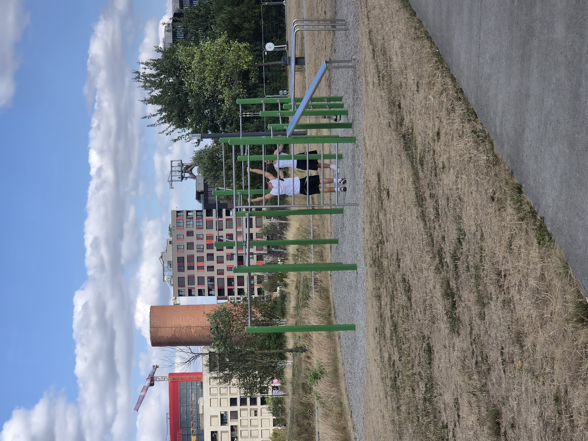 The image shows an outdoor gym area in the city of Sanem, Luxembourg. In the foreground, dry, yellow grass covers the ground. A paved pathway runs diagonally from the bottom right corner. In the mid-ground, a set of green-legged monkey bars with metal bars is visible. Two individuals, both wearing white t-shirts and dark shorts, are exercising on the monkey bars. One is hanging from a bar with their arms extended upwards, while the other is standing and reaching towards a bar. Behind the gym equipment, there is a gravel-covered area and more dry grass. The background features several buildings of varying architectural styles, including a tall, red cylindrical structure and modern apartment buildings with red window accents. A construction crane is visible to the left of the cylindrical building. Trees are scattered throughout the scene, and a cloudy blue sky dominates the upper portion of the image. Fencing and a round sign are visible in the far right background.