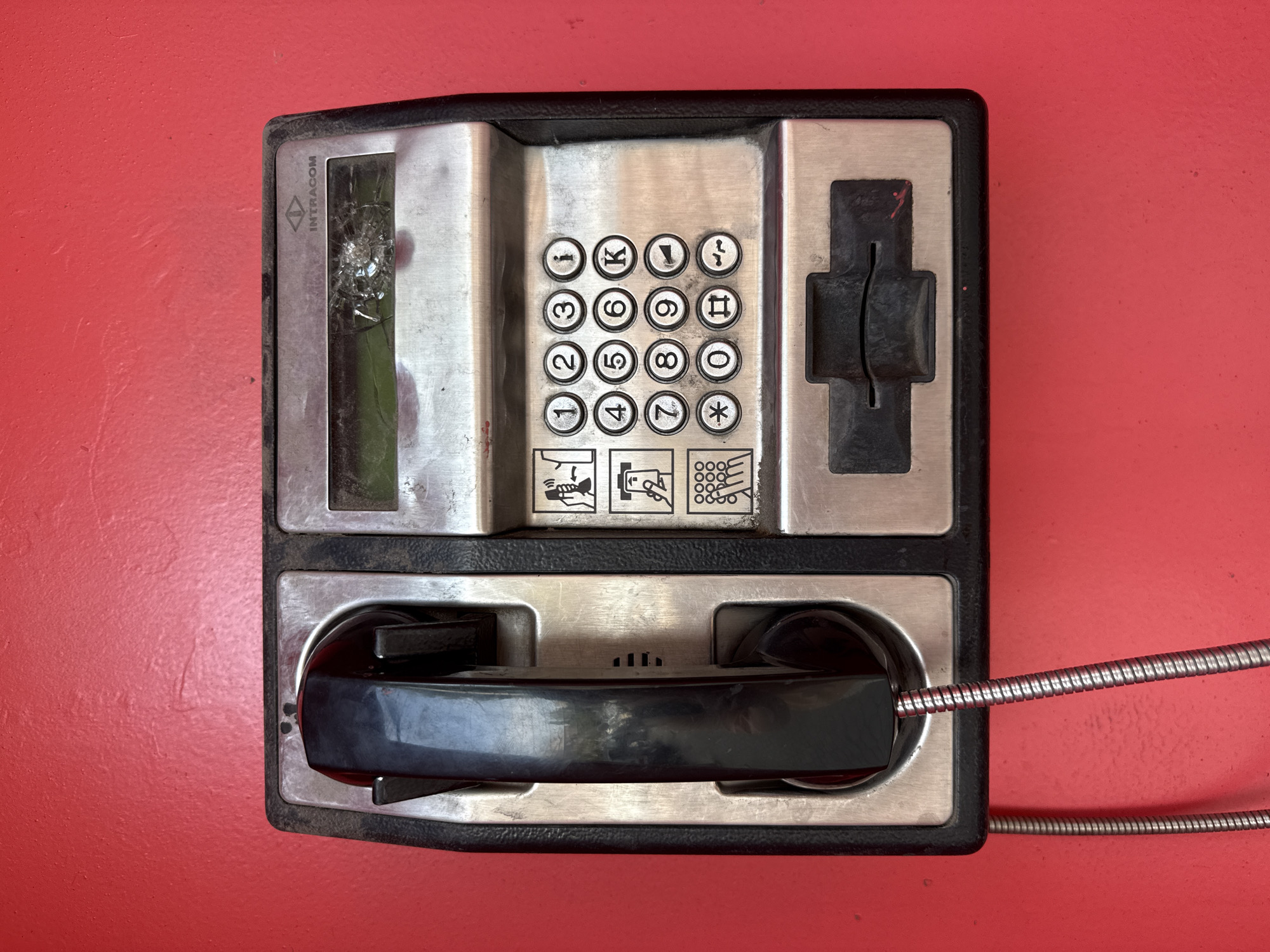 A public payphone is mounted on a vibrant red wall in Yerevan, Armenia. The payphone is divided into two main sections. The left section holds a black, coiled handset. The right section contains a cracked glass display screen at the top, displaying the "Intracom" logo. Below the screen is a keypad with numbers 0-9, along with symbols such as an asterisk and a hashtag. There are also icons above the keypad, one depicting a handset with sound waves, and another showing a person speaking into a microphone. A slot for coins or cards is located at the bottom of the right section. The payphone is made of metal and black plastic. The red wall shows some texture and a few minor imperfections.