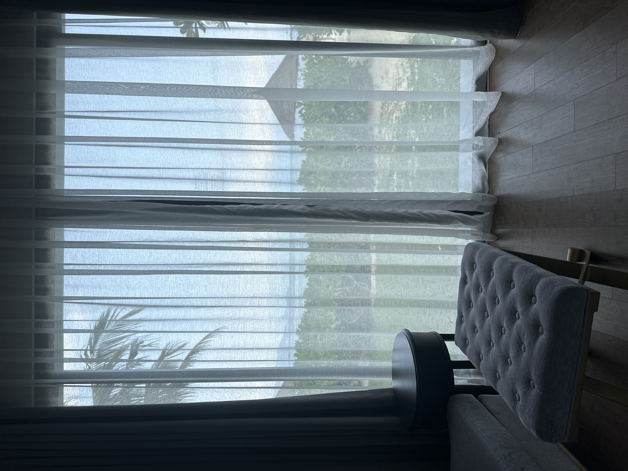 The image presents an interior view with a partially visible armchair positioned in front of a window. The window is covered with sheer curtains, allowing a filtered view of an exterior scene which appears to include a beach, water, vegetation, and potentially a structure in the distance. The room has wooden flooring. The lower portion of the image is darker, suggesting the presence of additional furniture or architectural elements contributing to a shadowing effect. The location is unspecified.