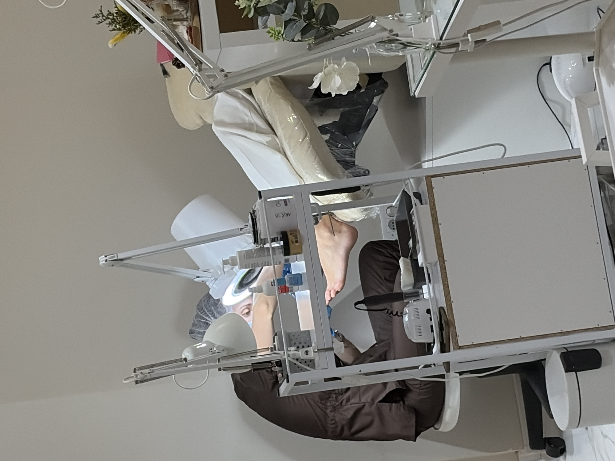 In Budva, Montenegro, a person is undergoing a pedicure. The individual is seated in a salon chair, with their foot resting on a raised platform. A nail technician, wearing a brown uniform and a hairnet, is meticulously working on the client's toenails. The technician is leaning over, illuminated by a bright magnifying lamp. Various tools and bottles of nail polish are arranged on a nearby cart, which is equipped with drawers and shelves. A white massage table is visible in the background, partially covered with a white sheet. The room appears to be a professional nail salon, with sterile equipment and a clean, well-lit environment.