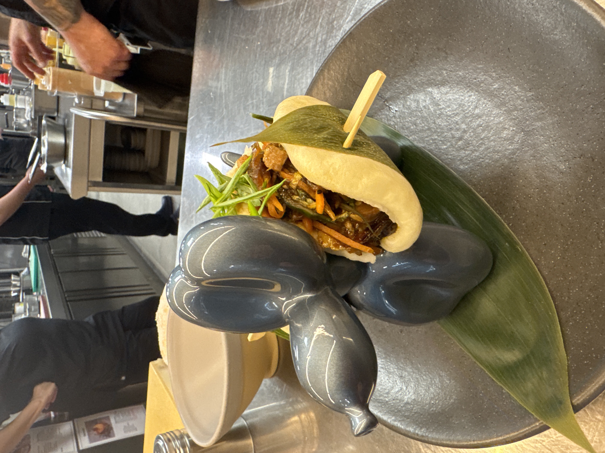 The image shows a food dish featuring a folded bun-like pastry filled with cooked vegetables and garnished with fresh greens. A wooden skewer pierces the bun. The dish is served on a large, dark-grey plate that includes a long, green leaf. A decorative grey sculptural form that looks like a balloon animal sits next to the bun. The plate is on a stainless steel surface. In the blurred background, a person in black clothing is working in what appears to be a kitchen area with stainless steel equipment. There is no explicit indication of location in Sofia, Bulgaria.