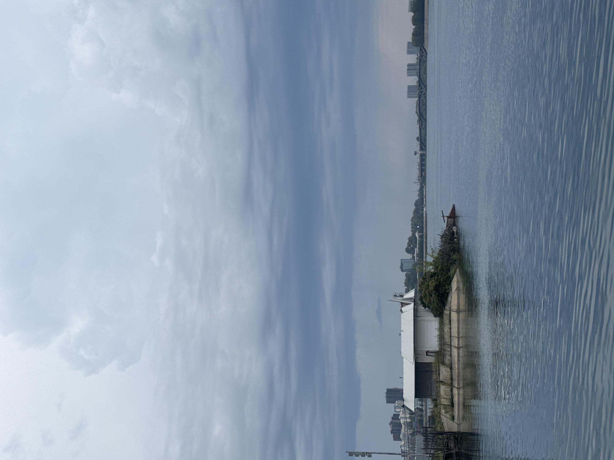 The image depicts a wide body of water under a cloudy sky, with a small island in the foreground. On the island, there is a white building with a peaked roof, surrounded by vegetation. In the background, a cityscape with several tall buildings and a bridge is visible across the water. The scene appears to be located in Irkutsk, Russia, likely along the Angara River or Lake Baikal, given the presence of a bridge and urban development characteristic of the city. The overall atmosphere is overcast and somber, suggesting a cool or possibly rainy day. There are no people visible in the image. The water exhibits ripples, indicating a slight breeze.