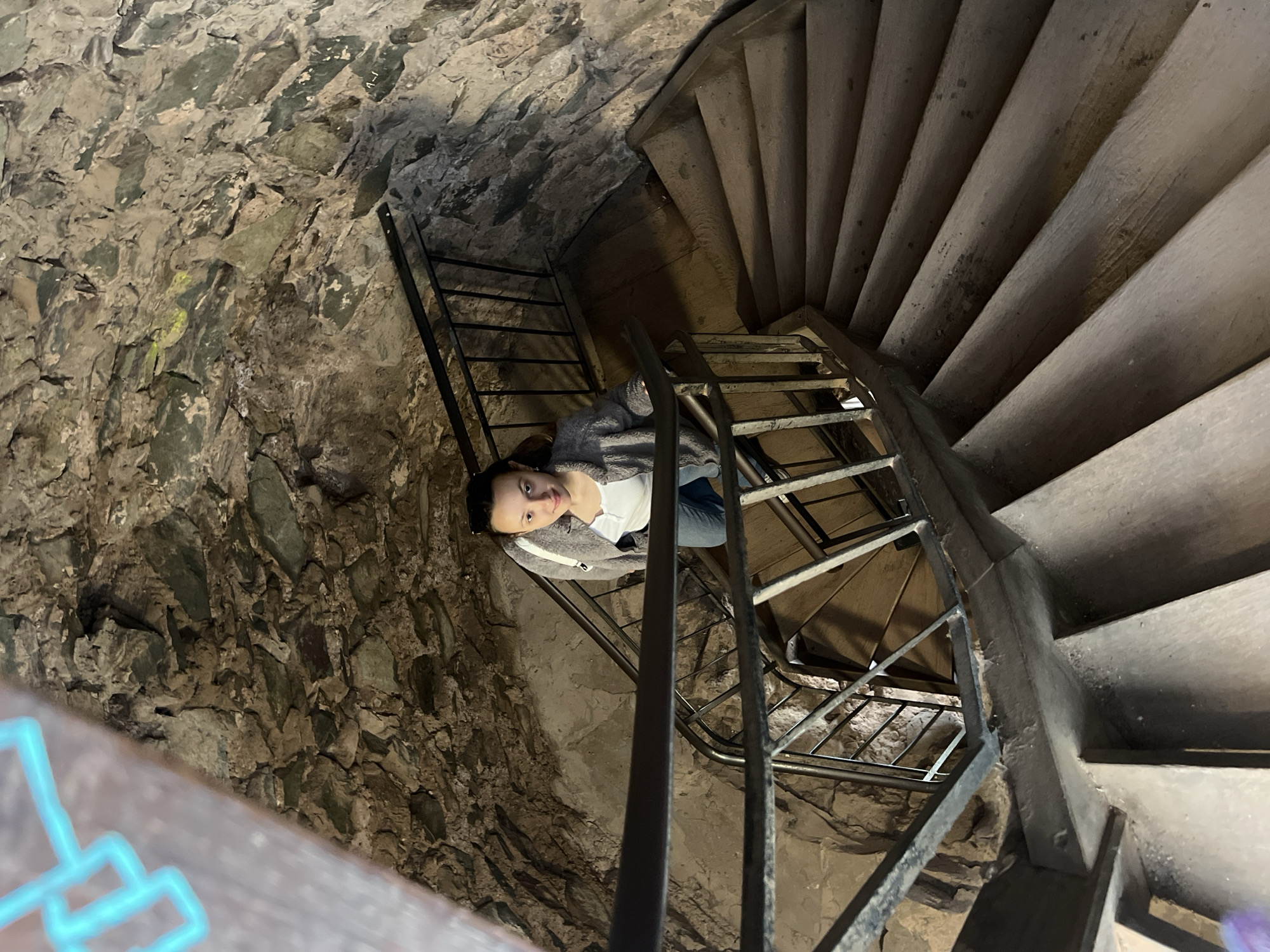 The image shows a woman on a spiral staircase in Saarburg, Germany. The staircase is constructed of stone and wood, with metal railings. The walls are made of rough-hewn stone. The woman is wearing a light-colored shirt, jeans, and a gray sweater. She is looking towards the camera. The setting appears to be an interior of a historic building, likely a castle tower due to the architecture.