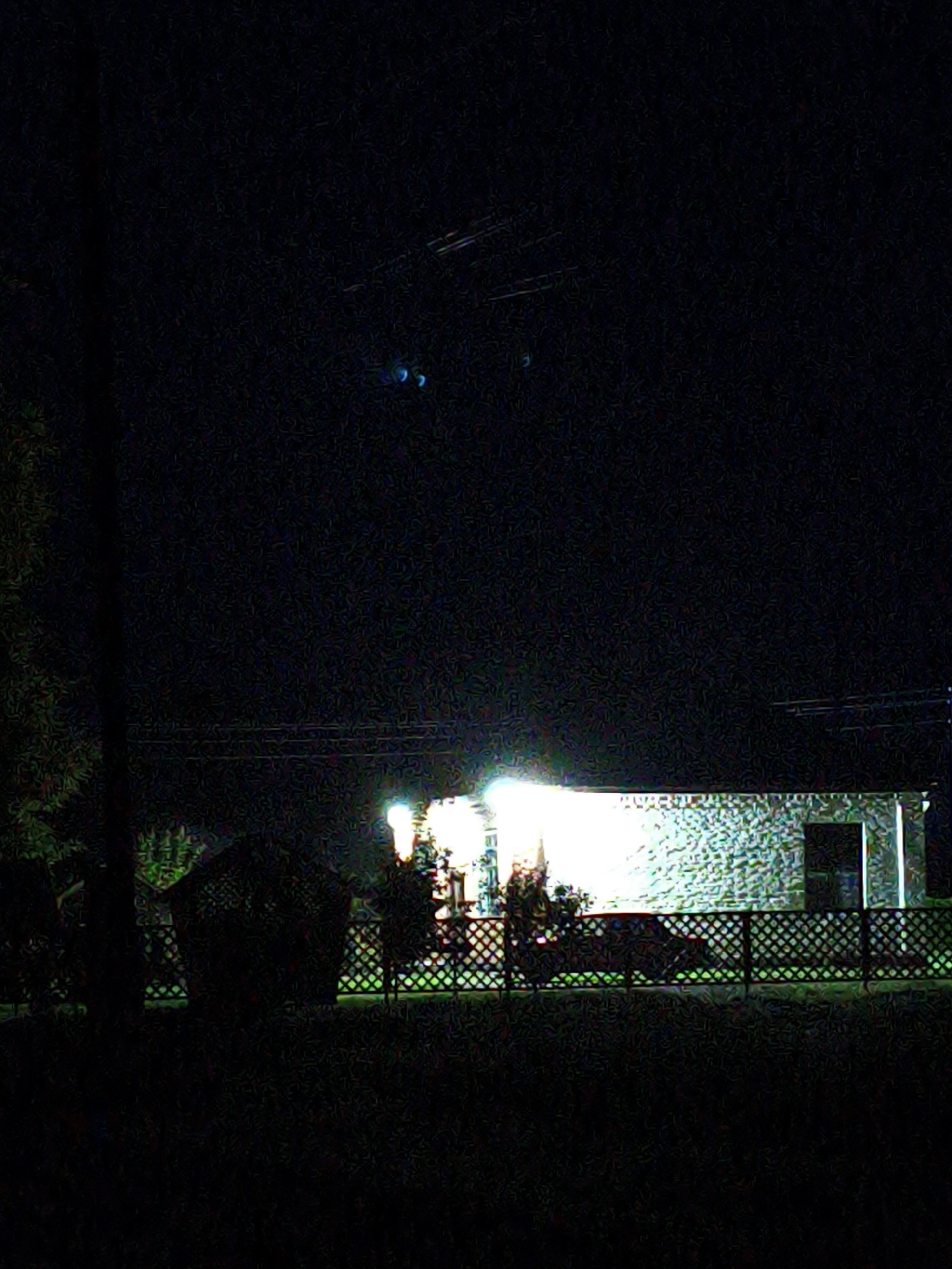 The image depicts a dark, outdoor scene at night, likely in Ustyvytsia, Ukraine. A building with a light-colored, textured facade is illuminated from within or by exterior lights, casting a bright glow. The building appears to have at least one dark doorway or window on its right side. In front of the building, there is a lattice fence with a diamond pattern, through which a green area is visible. To the left of the building, a dark, presumably low-lying structure, possibly a shed or gazebo, is silhouetted against the fence. Trees with dark foliage are present on the far left, obscuring the view of a vertical pole or utility pole. In the upper portion of the image, two distinct blue lights are visible against the dark sky, along with faint horizontal lines suggesting overhead wires or structures. The foreground is dark and appears to be a grassy or shrub-covered area. The overall image quality is grainy, suggesting it was taken in low light conditions.