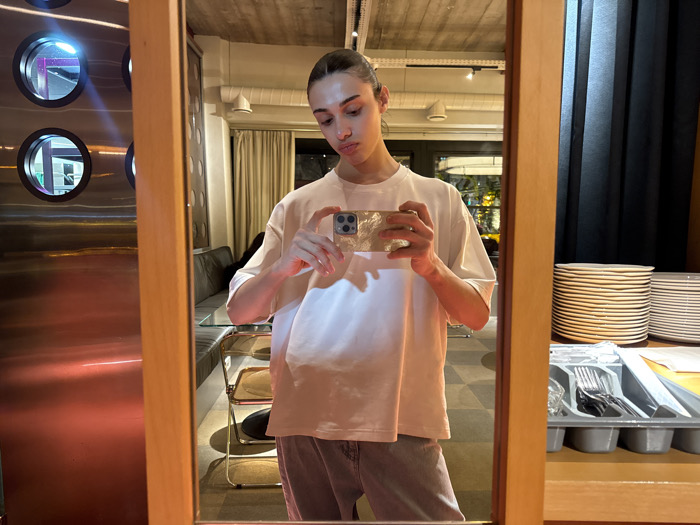 The image depicts an individual taking a mirror selfie with a smartphone in a modern interior setting, located in Yerevan, Armenia. The subject, a person with dark hair pulled back, wears a loose white short-sleeved t-shirt and light-colored trousers. They hold a silver-colored smartphone, featuring multiple rear cameras, horizontally at chest level, pointed towards the rectangular, light-wood framed mirror.
The reflection reveals various elements of the establishment. To the right, a tall stack of white ceramic plates and a smaller stack are visible on a dark counter, alongside a grey plastic tray filled with metal cutlery. A dark blue or black curtain hangs in the background. To the left, a section of a dark upholstered bench, a glass-topped table, and a chair with a golden metallic frame and dark seat are visible. The ceiling is exposed concrete with linear track lighting.
Behind the actual mirror, to the left of the reflected scene, a reflective metallic panel is present, featuring three circular apertures that capture and display internal light sources, including vibrant blue-purple and white illumination. The metallic surface itself shows a distinct orange-red glow from an unseen light source. The environment suggests a contemporary cafe, restaurant, or hotel dining area.