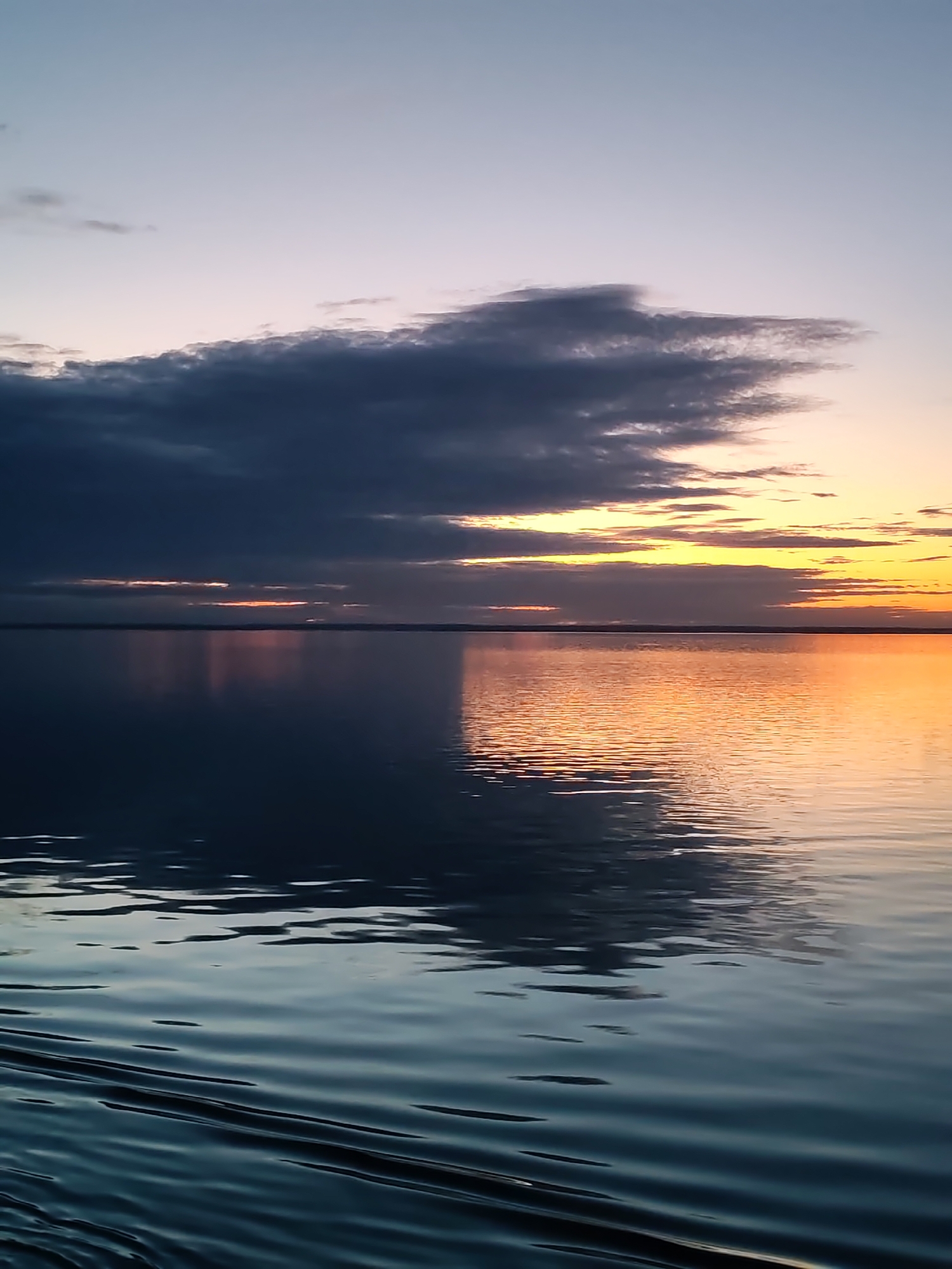The image captures a serene waterscape at either sunrise or sunset, presenting a vast expanse of water under a dramatic sky. The sky transitions from a soft, pale blue-grey at the top, descending into a large, dark grey cloud formation occupying the upper left quadrant. Towards the right horizon, streaks of vibrant orange and yellow light are visible, indicating the position of the sun, partially obscured by clouds.
The calm surface of the water acts as a mirror, perfectly reflecting the colors and formations of the sky. The reflection of the dark cloud mass appears in deep blues and greys on the left side of the water. Conversely, the right side of the water is bathed in a warm, glowing orange and yellow, mirroring the sun's light. A thin, dark line on the horizon separates the sky from the water, suggesting a distant landmass or coastline.
In the foreground, gentle ripples and waves create textured patterns on the water's surface, adding depth and movement to the otherwise still scene. A distinct V-shaped wake pattern is visible in the bottom left corner, hinting at recent movement on the water, though no boats or other subjects are present. The overall atmosphere is one of tranquil beauty, showcasing the interplay of light and shadow, and the rich color palette of dusk or dawn. No visible text is present.