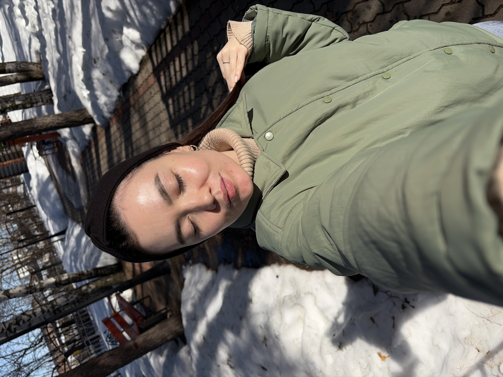 A young adult female is depicted in a close-up selfie, taken outdoors in Chaikovsky, Russia. She is positioned from the chest up, with her eyes closed, head slightly tilted, and a subtle smile. She wears a black headband, a beige turtleneck sweater, and an olive green collared jacket secured with light green buttons. A gold-colored hoop earring is visible in her left ear, and a ring is present on her right ring finger. Her right hand is raised, with fingers lightly touching her hair near her right shoulder. The background features a brick-patterned paved pathway bordered by remnants of snow. Bare deciduous trees, including birches, line the path, and a red park bench is visible in the mid-ground to the left. Distant architectural structures are faintly discernible through the trees. The scene is brightly illuminated by sunlight, casting distinct shadows. The environment indicates a winter or early spring day.