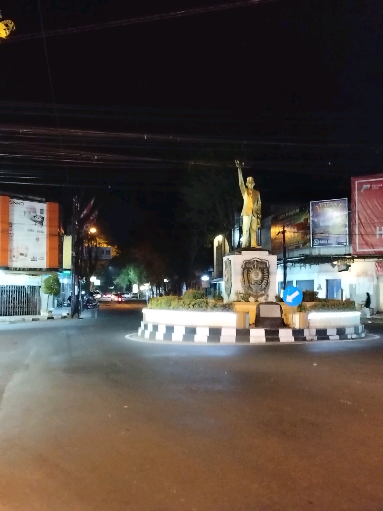 This is a nighttime photograph taken at the Alun-Alun Purworejo, a public square in Purworejo, Indonesia. The focal point of the image is a statue of a man in a suit, with his right arm raised. The statue is situated on a tiered pedestal adorned with decorative elements and a crest. Around the base of the statue is a circular planter with white and black checkered borders. A blue circular traffic sign with a white arrow indicating a left turn is visible on the planter.
The statue is located in the center of a roundabout. The road surface is asphalt. In the background, there are several buildings and billboards, some illuminated. Trees line one side of the street. Streetlights cast a warm glow on the scene. There are a few vehicles, including cars and motorcycles, visible on the road. A person is walking on the sidewalk in the background. The sky is dark, indicating it is night. The overall impression is of a public space at night, with the statue as a prominent landmark.