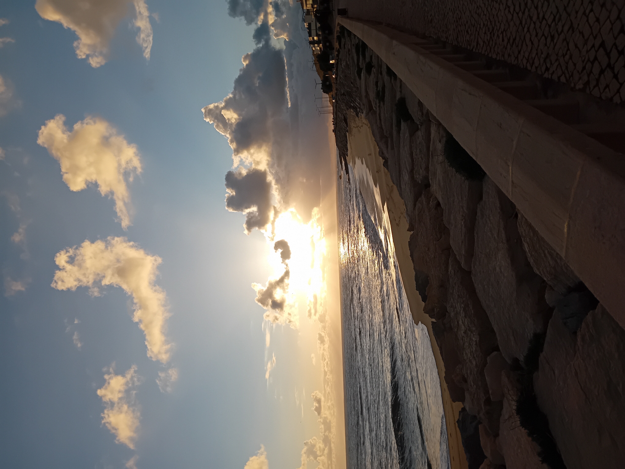 The image depicts a sunset over the ocean in an unidentified city. The sun is partially obscured by clouds, casting a golden glow on the water and sky. A stone embankment runs along the edge of the beach, with a cobblestone walkway above it. The image has been rotated 90 degrees counter-clockwise. Wind turbines are visible along the horizon.