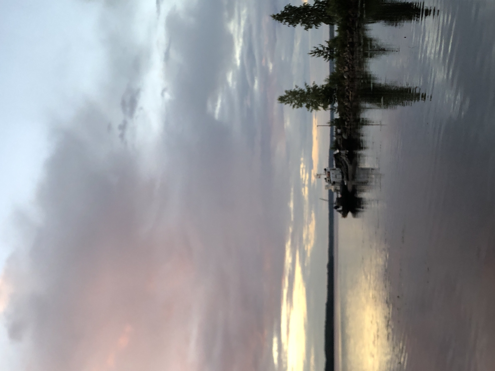 The image depicts a serene waterfront scene at dusk, possibly in the city of Svirstroy, Russia. A calm body of water, likely a river or lake, dominates the foreground and middle ground, reflecting the soft hues of the twilight sky. The sky is filled with dramatic clouds, painted in shades of pink, purple, and pale blue, suggesting the last moments of daylight.
Along the distant shoreline, a line of trees forms a dark silhouette against the lighter sky. In the middle ground, near the shore, several boats are docked. The largest boat appears to be a white vessel with some superstructure. To its right, smaller, darker boats are moored. The shoreline itself seems to be constructed with rocks and possibly some vegetation.
The overall atmosphere is peaceful and tranquil, capturing the natural beauty of the environment during the transition from day to night. The reflections on the water add to the sense of calm and create a mirror image of the sky and the objects on the shore. There are no visible people in the image.