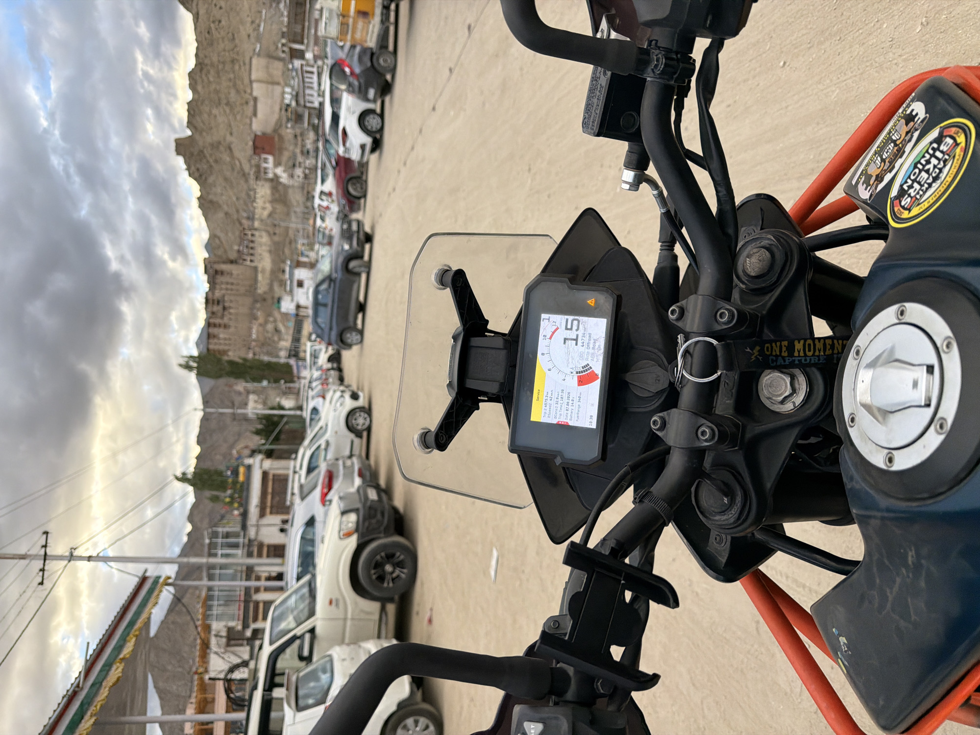 The image is a first-person perspective shot from a motorcycle in Leh, India, looking towards a parking lot filled with vehicles and a mountainous landscape with buildings in the background. The motorcycle's dashboard displays a speed of 15 km/h, trip information, and the date "07/07/2026." Several cars, including SUVs and hatchbacks, are parked on a dirt surface. The background features a town with traditional architecture and a prominent fortress or palace situated on a hill. The sky is overcast with dramatic clouds. The scene appears to be a common tourist area, possibly near a checkpoint or viewpoint. There are no people visible in the frame.