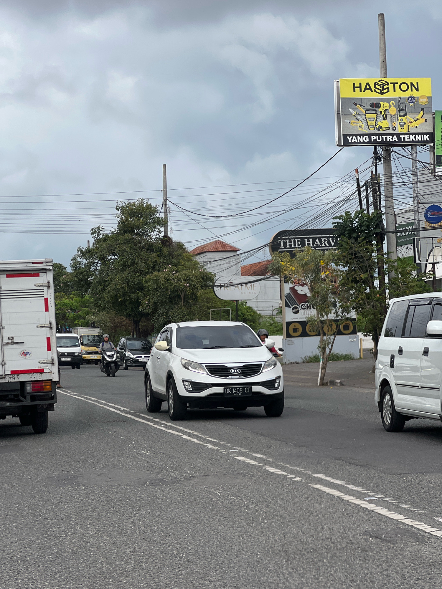 The image shows a street scene in Denpasar, Indonesia. The sky is overcast with a grey, cloudy sky. Power lines are visible crisscrossing the frame. On the right side of the image, a large billboard displays "HASTON" in yellow lettering above images of power tools. Below this, it reads "YANG PUTRA TEKNIK". Further down the street, another sign for "THE HARVEST" is visible, featuring a stylized image of food.
Several vehicles are on the road. In the foreground, a white Kia Sportage SUV is facing left, its license plate reading "DK 1408 CT". To its right, a white van is partially visible, facing left. A white truck is on the far left of the image, facing left. In the middle ground, a person is riding a motorcycle, wearing a grey helmet and jacket, facing towards the viewer. Other vehicles, including a yellow truck and a black car, are further back on the road. Trees and buildings line the street. The road is asphalt and has white lane markings.