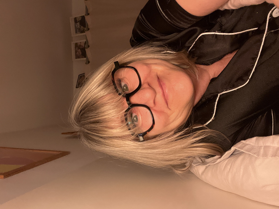 A woman with blonde hair, wearing black glasses and a black pajama-style top with white trim, reclines on a white pillow. Her face is angled towards the camera, and she has a faint smile. In the background, there is a cream-colored wall with small framed pictures. A portion of a picture frame sits on a ledge in the corner. The lighting appears soft and warm.