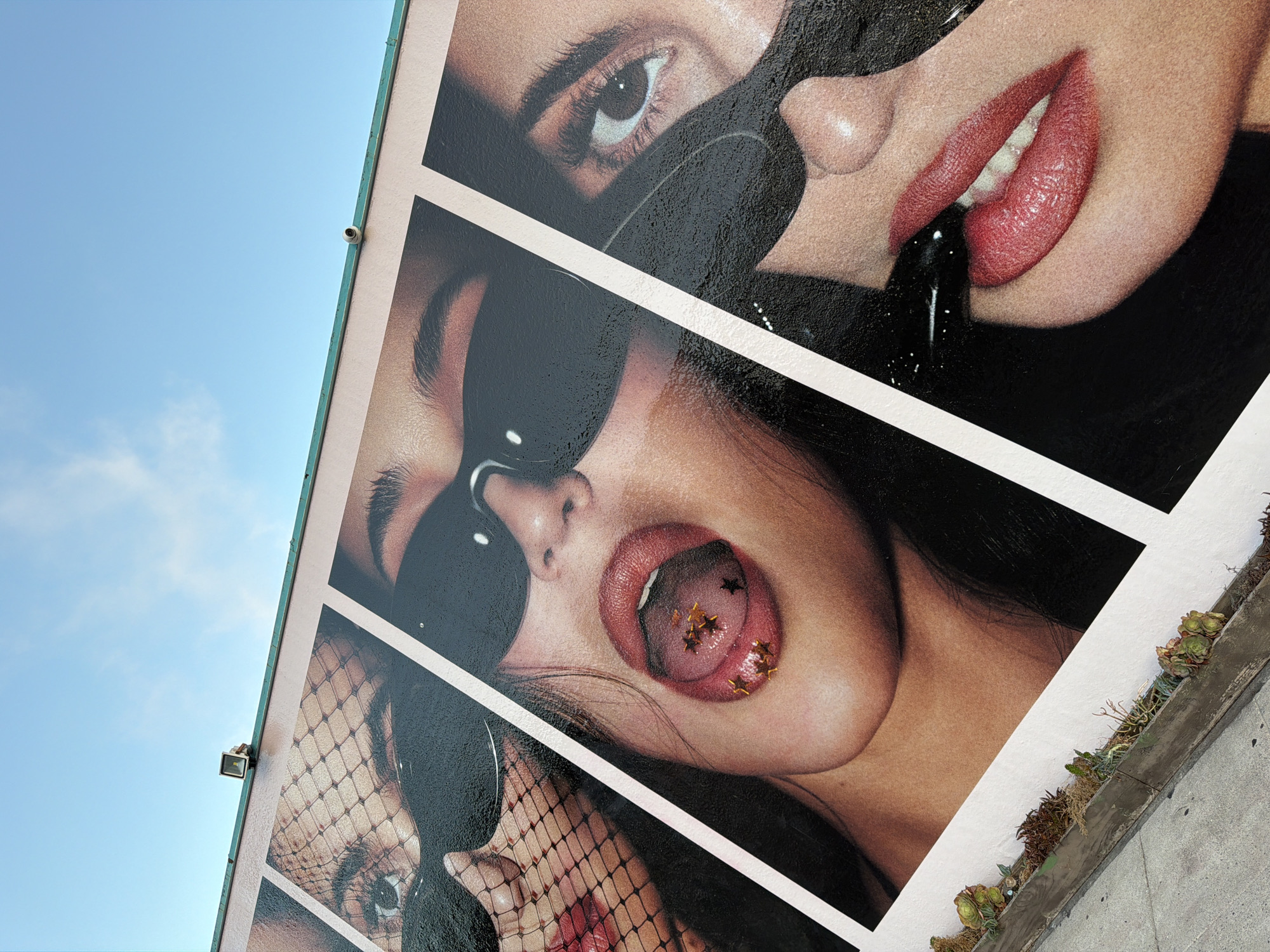A mural featuring a close-up of a woman's face is displayed on the exterior wall of a building. The mural is divided into panels, with the woman's features cropped and repeated. In one panel, her tongue, adorned with gold star-shaped glitter, is visible. In another, her lips are pursed around a black object, possibly a lipstick tube. The mural is positioned at an angle, suggesting it's on a sloped wall or a building corner. Above the mural, the sky is visible, with a few wispy clouds. Two small surveillance cameras are mounted on the roofline above the mural. In the foreground, a concrete sidewalk runs alongside a planter box filled with succulents and dry moss. The overall scene captures a vibrant, urban aesthetic, likely in the city of Los Angeles, United States of America.