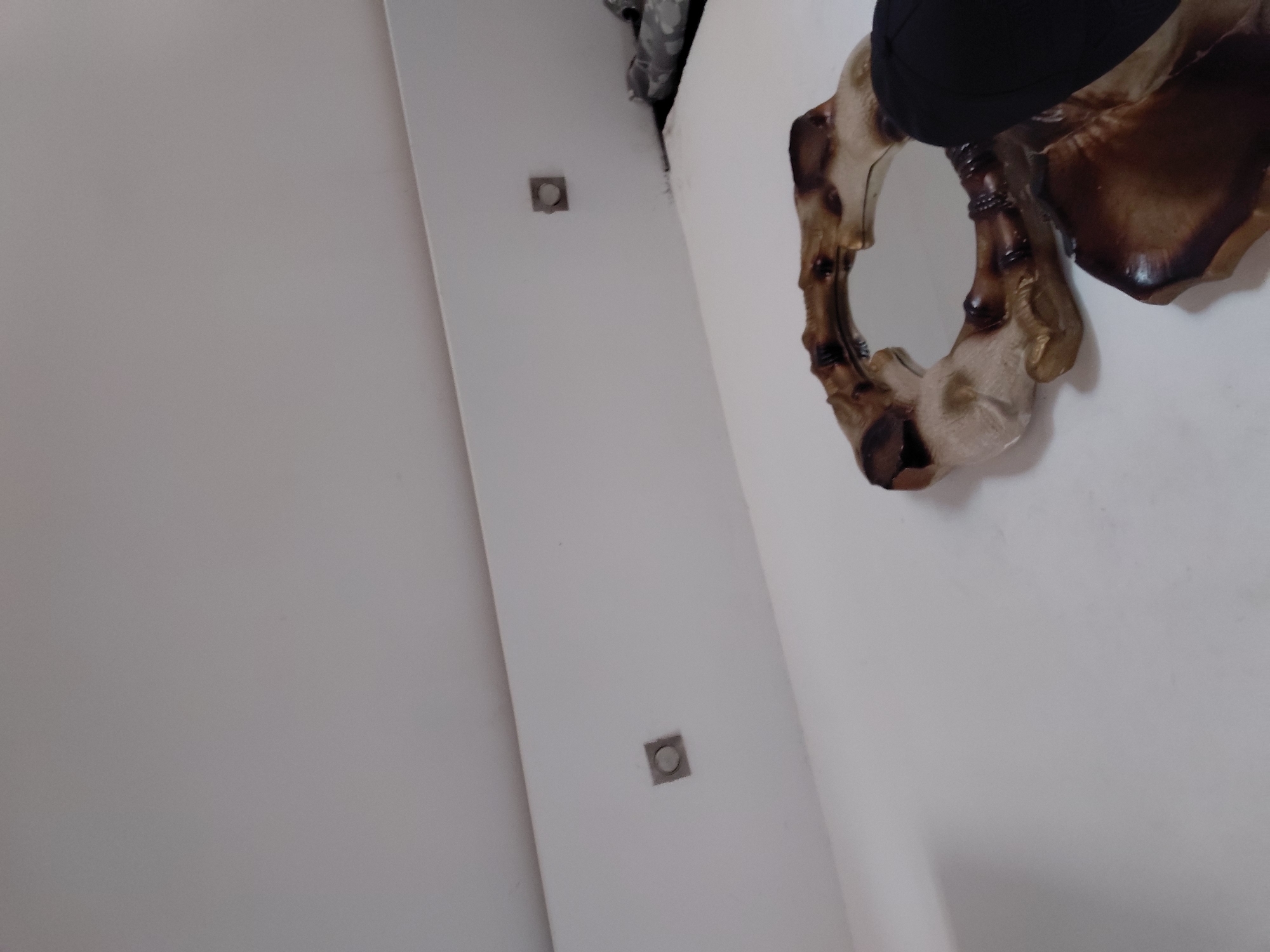The image displays an indoor scene dominated by a plain, light-colored wall, likely white or off-white. The perspective is angled, suggesting the wall might be a ceiling or a highly tilted surface.
In the upper right section, a prominent decorative object is visible. This object is a sculpture or an ornate mirror frame, fashioned from a material with a textured, organic appearance, resembling wood or bone. It features a striking contrast between darker, reddish-brown tones, some almost burnt or charred in appearance, and lighter, creamy beige sections. Its intricate form is abstract, with curves and hollows, somewhat evoking a stylized anatomical structure or a piece of natural art. Resting on top of this decorative piece is a solid black object, possibly a cap, fabric item, or part of clothing.
Further to the left on the wall, two small, square, recessed fixtures are present. Each fixture has a metallic-looking border and contains a circular element in its center, suggesting they could be flush-mounted lights or decorative wall inserts.
The setting is an interior space, with no people present and no visible text. The lighting appears consistent with an indoor environment. Given the location context, this scene is situated in Santa Ana, Venezuela, though no specific visual cues within the image confirm this geographical detail. The overall impression is of a static display or part of a room's decor.