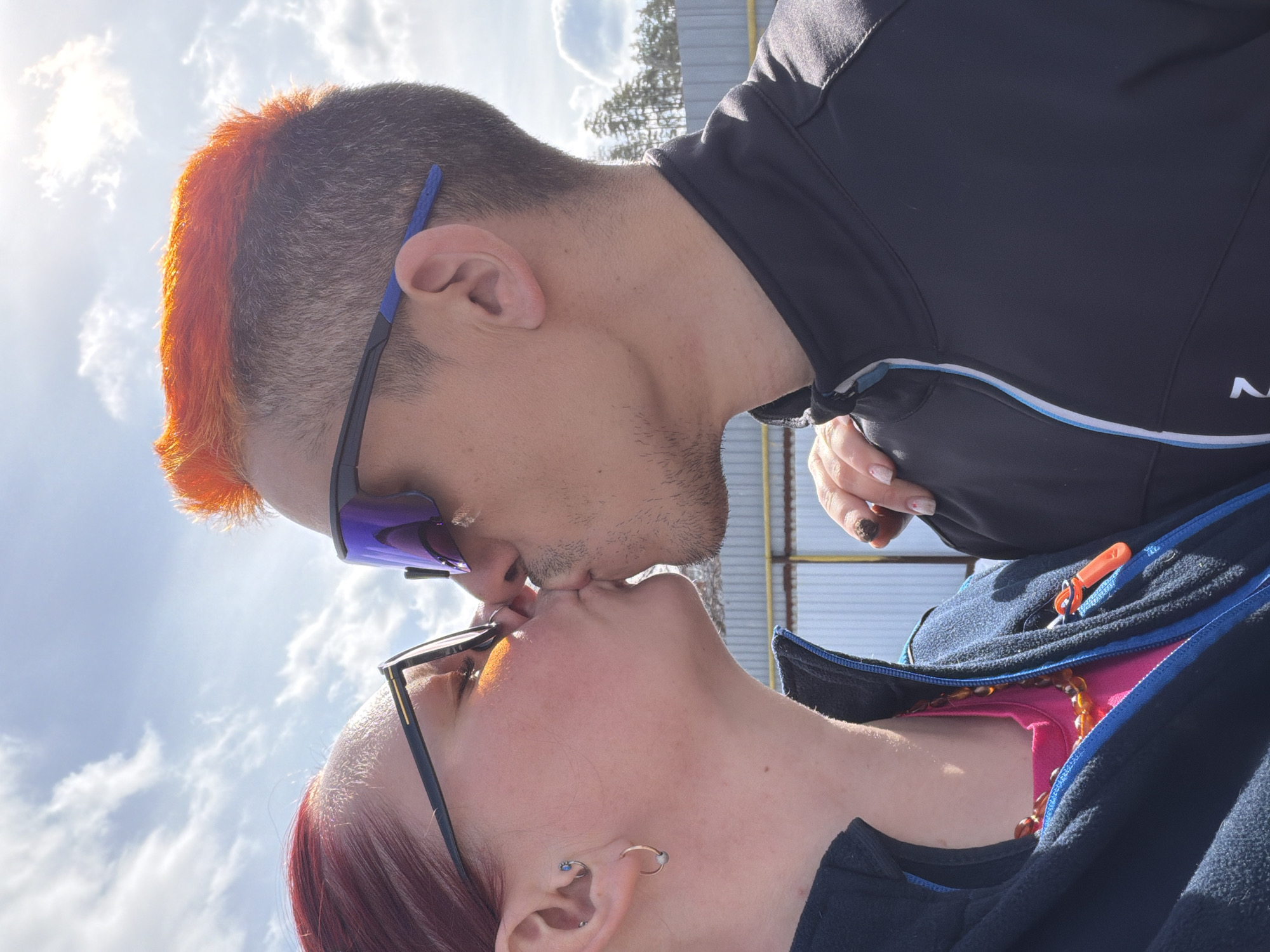 A couple, a male and a female, are kissing. The male has dark hair with orange highlights, wears sunglasses, and is wearing a black top. The female has red hair and wears glasses. She is wearing a blue jacket over a pink top, along with a necklace. The background shows a cloudy sky and a building structure. The location is likely Chaikovsky, Russia.