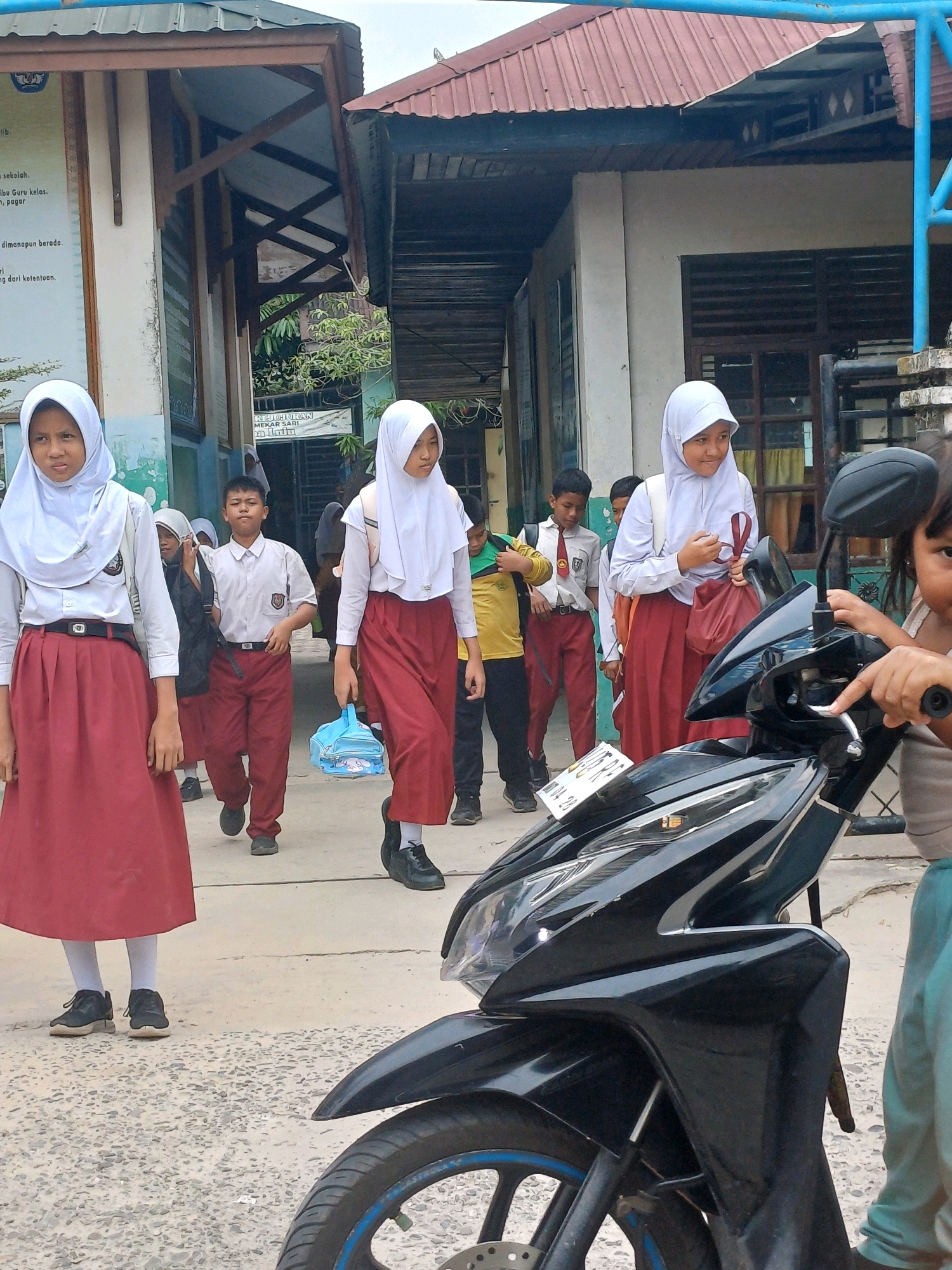 In Dumai City, Indonesia, a group of schoolchildren in uniform are walking outside on a paved area near a building. The children, mostly wearing white hijabs and red skirts or pants, are walking towards the viewer. One girl in the foreground on the left is wearing a white hijab, a white shirt, and a red skirt, with black shoes. To her right, a boy in a white shirt and red pants is walking, carrying a black backpack. Another girl on the right, also wearing a white hijab, white shirt, and red skirt, is holding a light blue bag. Further in the background, other children in similar uniforms are visible. In the foreground, a black motorcycle is parked, with its front wheel and handlebars clearly visible. A young child's hand is reaching towards the motorcycle's handlebars. The environment suggests an outdoor area near a school, with a building with a corrugated metal roof visible in the background. The sky is overcast.