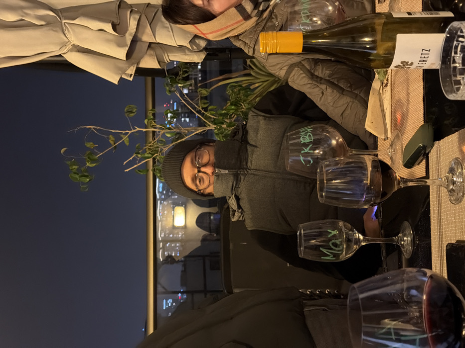 A group dines outdoors at night, a cityscape visible in the background. A person wearing a gray beanie and glasses is seated, with three wine glasses, two containing red wine, in front of them. One glass has the name "Max" written on it. Another person is partially visible wearing a scarf and a jacket. A bottle of white wine, labeled "Herretz" is also on the table.