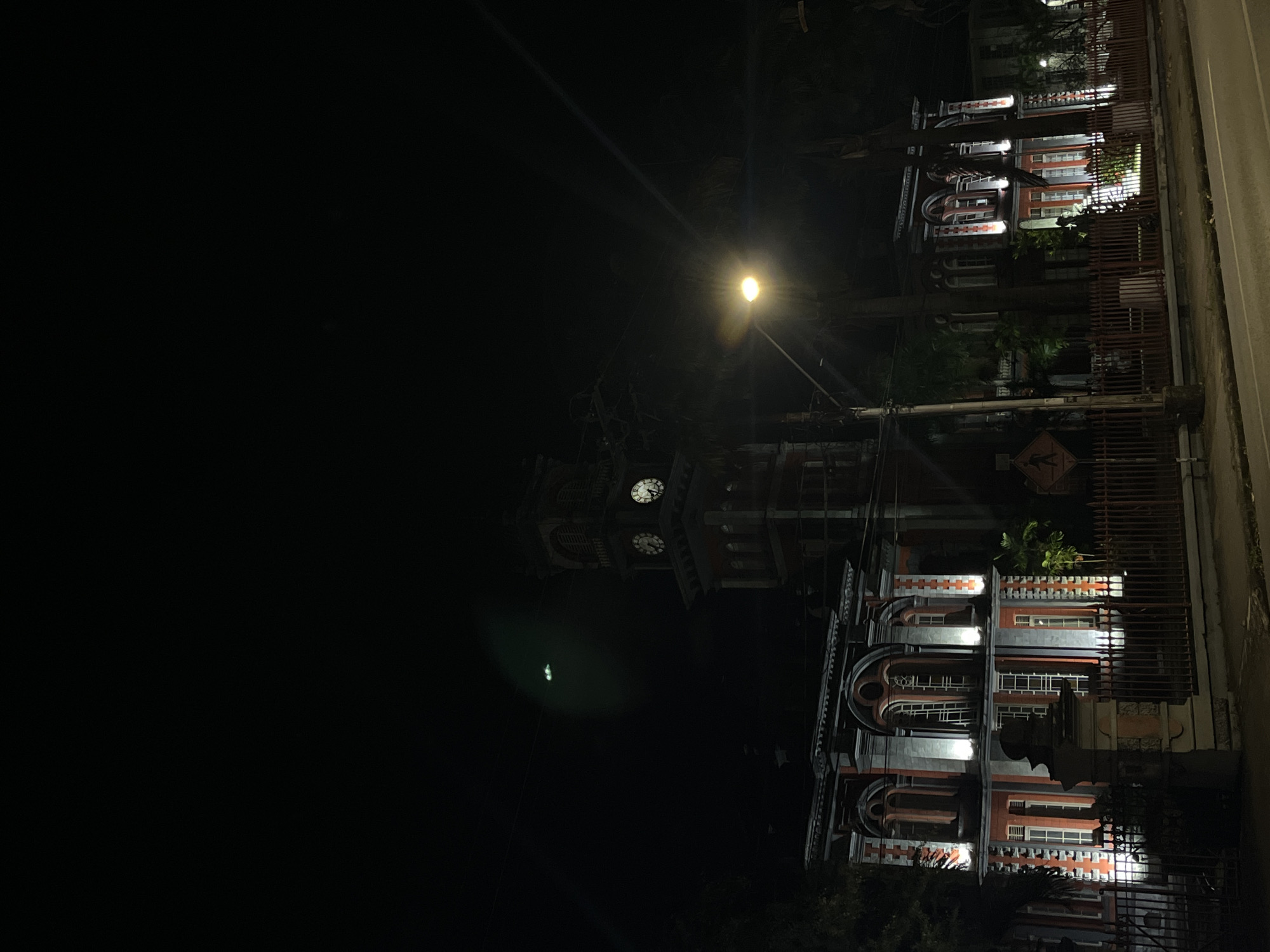 The image shows a building at night in Port of Spain, Trinidad and Tobago. The building has red and white trim and appears to have multiple floors. A street light is visible above the building, and there is a clock tower. The image is oriented sideways, with the building appearing on the right side of the frame. A fence is visible in the foreground. The night sky is dark and occupies the majority of the image. Based on the architecture and location, the building appears to be the Red House (Parliament Building).