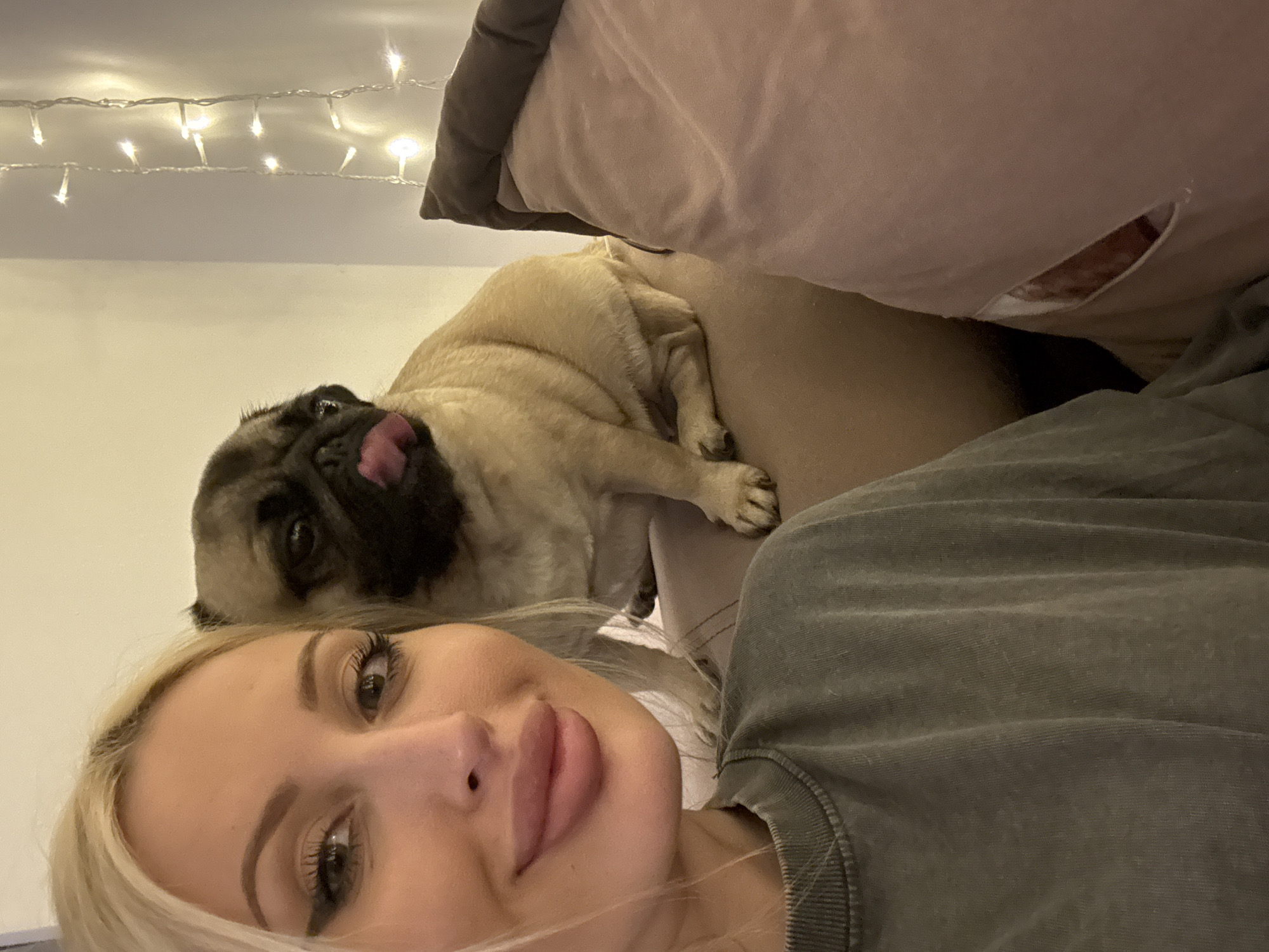 A medium shot captures a woman and a pug in an indoor setting. The woman, in the foreground and to the left, has fair skin, blonde hair, and is wearing a dark green t-shirt. She has noticeable makeup, including winged eyeliner and full lips. Her expression is friendly, with a slight smile. Behind her and to the right, a fawn-colored pug is seated. The pug is sticking its tongue out, its eyes are open, and it appears to be looking slightly to the right. The background features a plain wall with a string of fairy lights. A plush, light-pink pillow is visible to the far right. The overall lighting is soft and warm. The image's location is Unknown, Unknown.