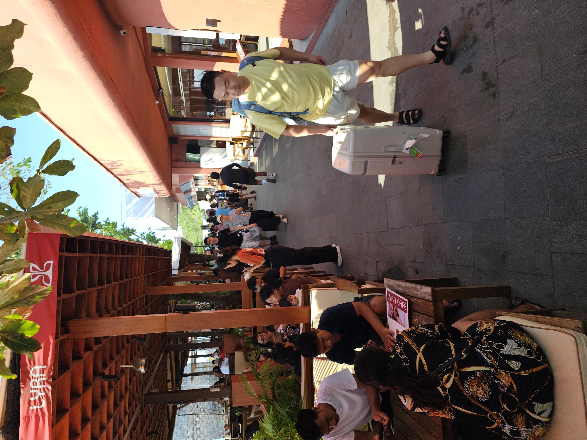 A man in a yellow t-shirt and light-colored shorts, wearing black sandals, is walking with a light-colored suitcase on wheels. He has a backpack on. He is walking on a tiled pathway in front of a building with an orange facade and glass windows. To his left, there is an outdoor seating area of a cafe with wooden tables and chairs. Several people are seated at the tables, some looking at their phones and others at menus. A woman in a black patterned dress is sitting at a table, her back to the camera. The cafe has a wooden roof structure overhead with a red banner that reads "LYRA" in white lettering. The scene appears to be in Denpasar, Indonesia, likely a tourist area or commercial street.