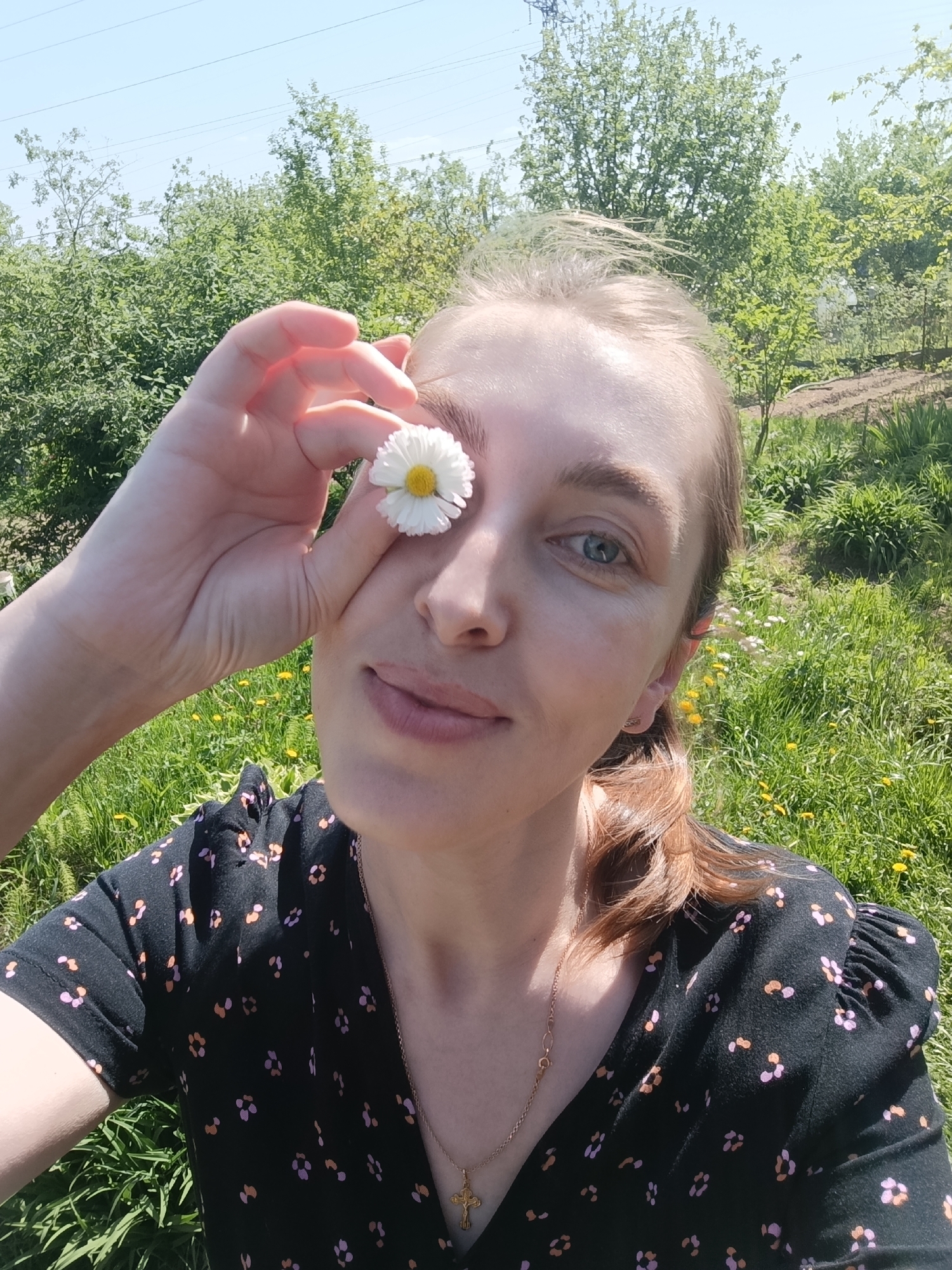 A woman is taking a selfie outdoors, holding a daisy in front of one eye. She has light skin, light brown hair pulled back, and is wearing a dark floral dress with a gold necklace. The background shows green grass, trees, and a clear blue sky. The location appears to be a rural or garden area. It is impossible to determine if the location is in the city of Chaikovsky, Russia, from the provided image.